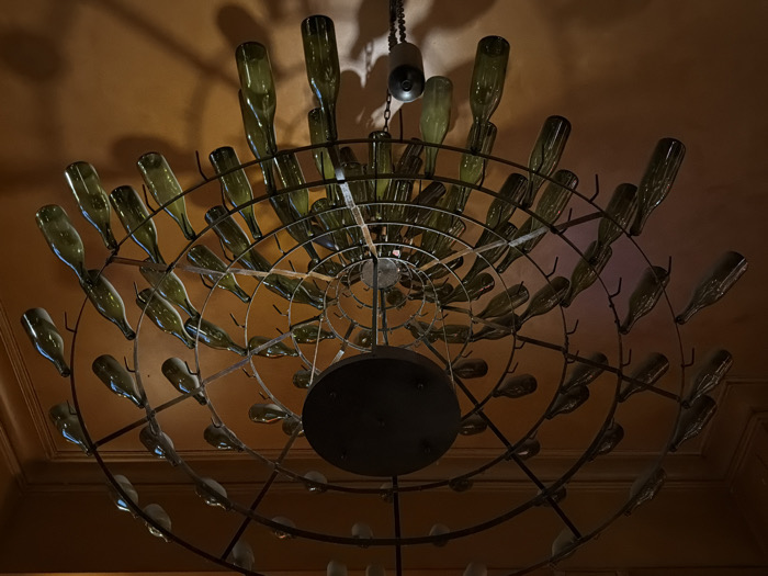A low-angle shot reveals a circular chandelier constructed from metal rings and numerous repurposed green glass bottles. The bottles are arranged in concentric circles, suspended upside down and radiating outward from a central point. A dark ceiling is visible behind the fixture, illuminated by the light emanating from it. A chain hangs from the ceiling, connecting the chandelier. Shadows from the bottles cast patterns on the surrounding surface, adding depth to the composition.
