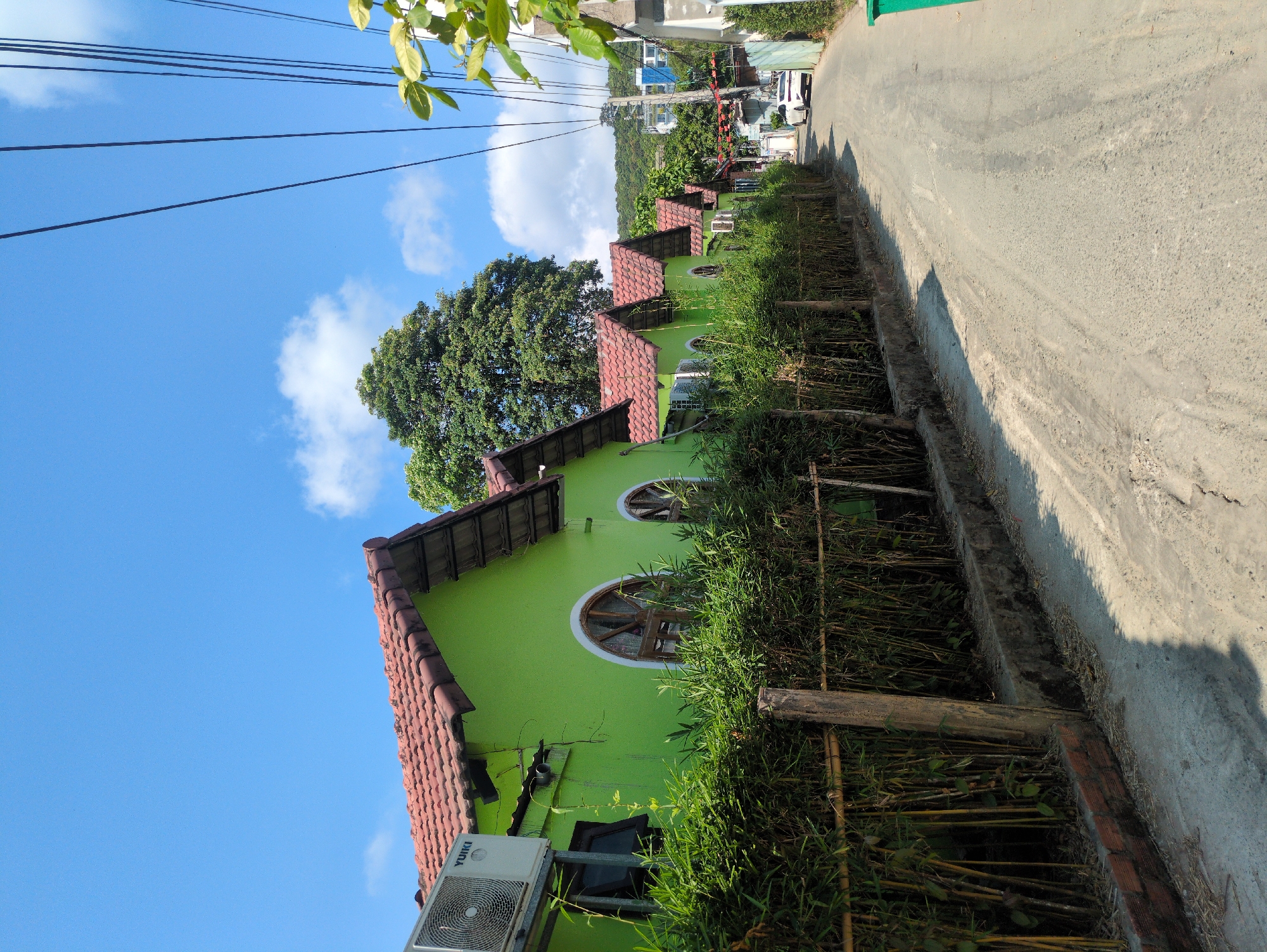 The scene is a street view in Phú Quốc, Vietnam, showing a row of bright green buildings with red tiled roofs along the edge of a paved road. The buildings are lined with bamboo fencing and dense greenery. An air conditioning unit is visible on one building. Above the buildings, power lines stretch across a partly cloudy blue sky. Further down the road, other buildings and parked vehicles can be seen. The angle of the photo is tilted, making the street and buildings appear at an angle.
