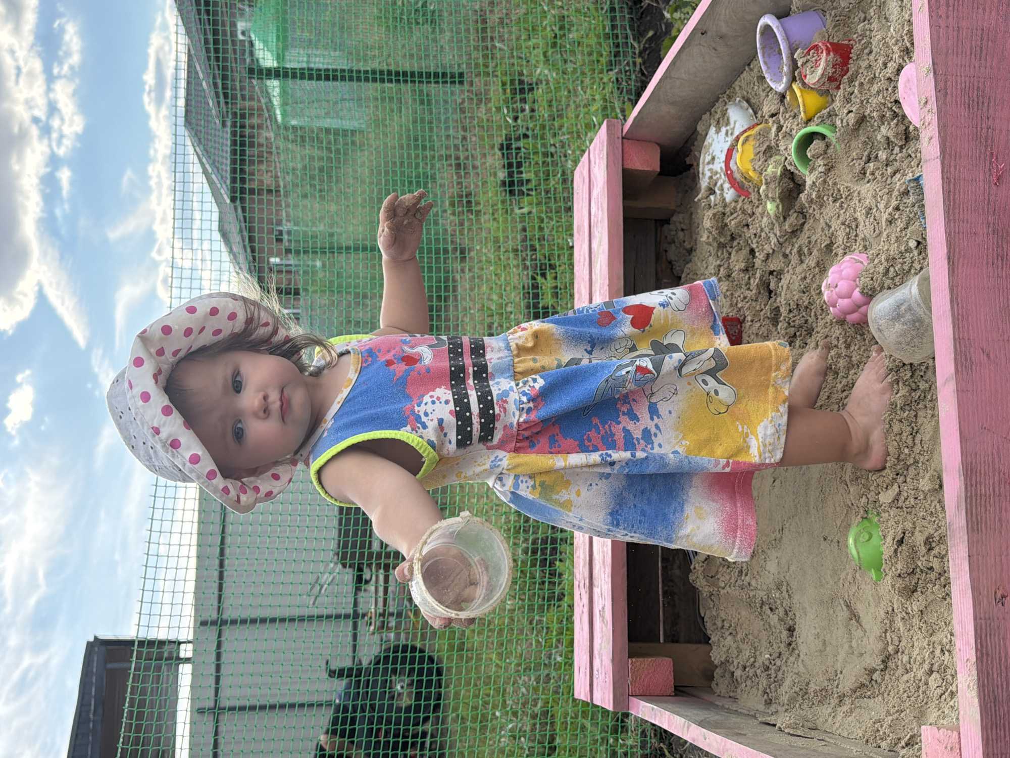 A young child, a girl, is playing in a sandbox. She is wearing a colorful dress with cartoon characters and a polka-dot hat. The sandbox is made of pink wood and is filled with sand and various toys, including buckets and molds. The child is holding a small plastic cup in her hand, and her feet are visible in the sand. The scene appears to be outdoors, with a green fence and a cloudy sky in the background. The overall impression is one of playful innocence and childhood enjoyment. The time of day is likely daytime, given the bright sky. The location is noted as Krasnyi Krym, Russia.
