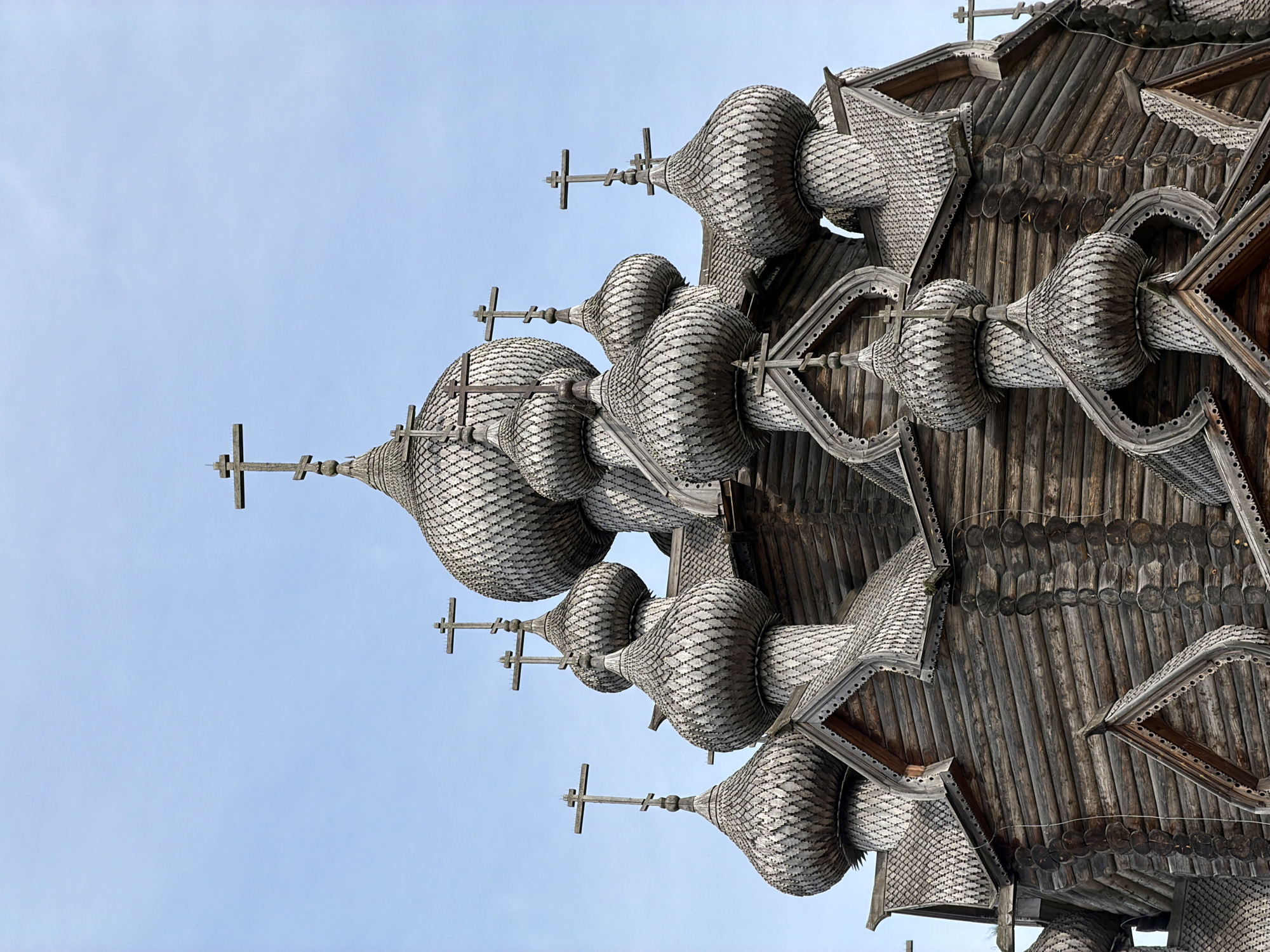 The image shows a close-up, eye-level view of the wooden domes and crosses of an Orthodox church in the city of Imeni Sverdlova, Russia. The architecture is traditional, with numerous onion-shaped domes covered in wooden shingles, each topped with a multi-barred cross. The church is constructed from logs, clearly visible in the stacked construction of the walls. The sky is a clear, pale blue, providing a simple backdrop to the intricate wooden details of the church. No people or other objects are visible in the frame.