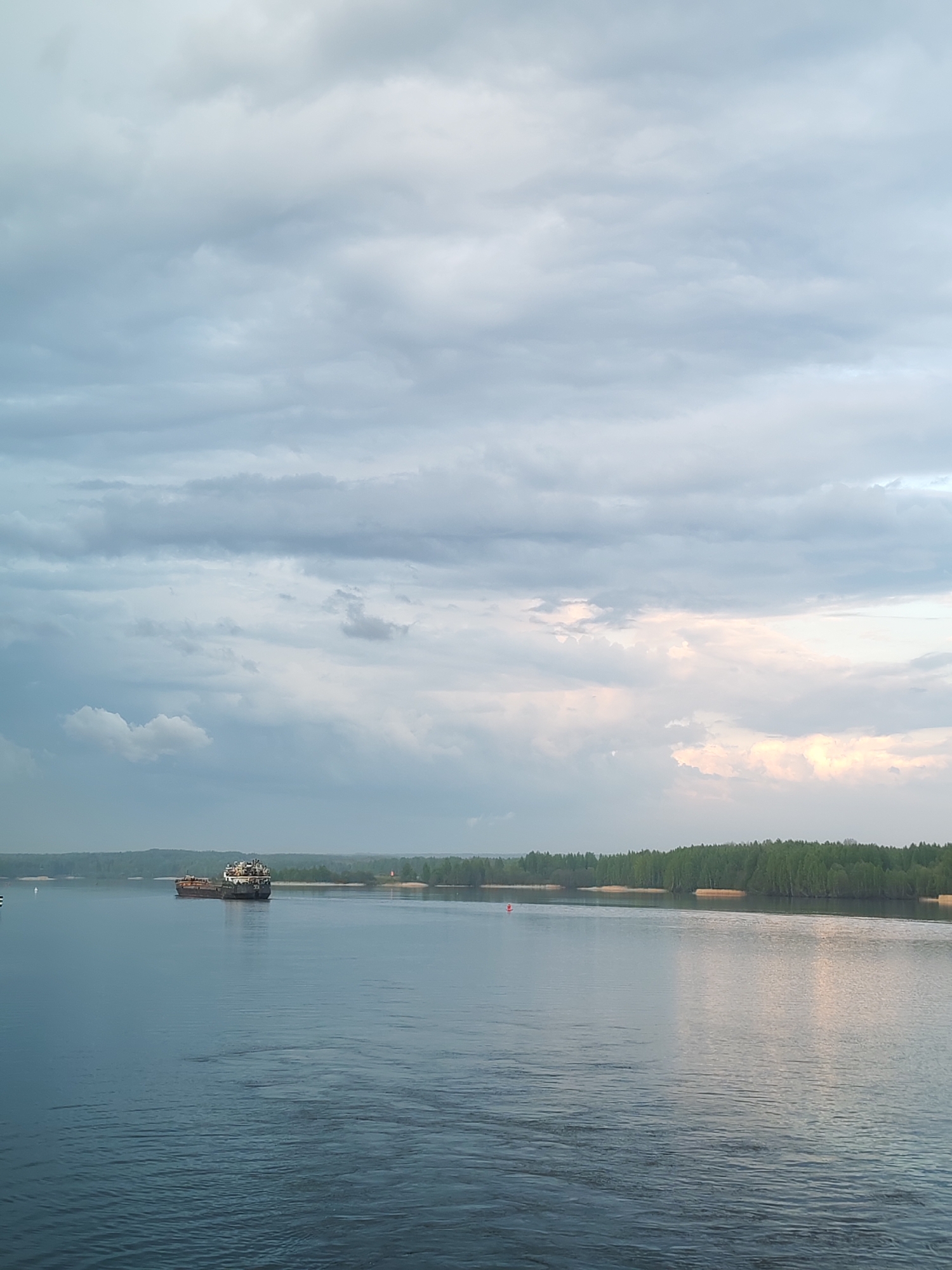 The image depicts a wide, calm body of water, likely a river or large lake, under a sky filled with layered, predominantly gray and blue clouds. There are patches of lighter, almost white clouds, suggesting a potential break in the overcast. A forested shoreline is visible in the distance, with a strip of light-colored sand or beach in front of the trees. A cargo ship is visible on the water. The overall setting gives the impression of a natural, possibly rural or suburban, environment. Ankhimovo, Russia is on the banks of the Volga. The photograph shows a waterway with the characteristics of the Volga river.