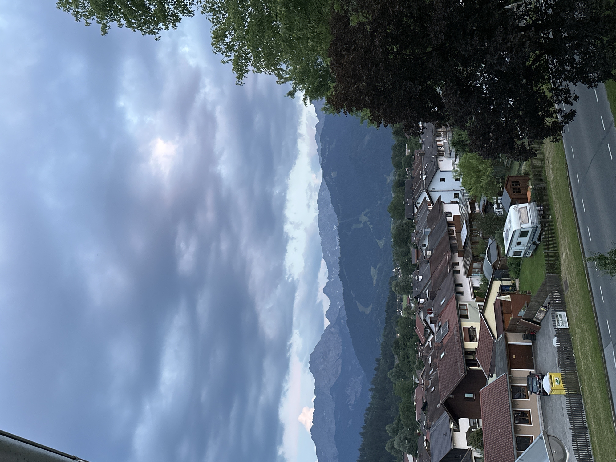 This media file is an outdoor, eye-level, landscape photograph taken from a high vantage point, likely a balcony or rooftop, looking out over a residential area and mountainous terrain. The orientation of the photo is such that the sky and mountains occupy the left and center portions, while the right side shows a street, houses, and trees.
The scene depicts a town nestled at the base of imposing mountains. The sky is dramatic, filled with a mix of dark, ominous clouds and patches of brighter light, suggesting either a pending storm or the aftermath of one. Sunlight is breaking through the clouds in the upper left quadrant, creating a diffused glow. The mountains in the background are layered, with the closest ones appearing dark green and densely forested, while the more distant peaks are a muted blue-gray and jagged, indicating rocky outcrops and significant elevation.
In the foreground and mid-ground, a cluster of houses with red-tiled roofs lines a street. The architectural style appears European, with some houses featuring multiple stories and balconies. The street itself is paved and has white lane markings. A few trees, some with dark foliage, are interspersed with the houses. Visible objects include a white caravan parked in a yard, a small wooden shed, and what appears to be a small construction vehicle with a yellow and white body near a garage. There is also a fence separating the residential properties from the road.
The time of day appears to be late afternoon or early evening, given the quality of the light and the dramatic cloud formations. The weather seems to be overcast and possibly humid, with the potential for rain.
There is visible text on a signpost in the lower right quadrant of the image. It reads "ZImmervariante STAMMER".
There are no discernible people engaged in specific activities, though one figure might be present near the construction vehicle. The overall emotion conveyed by the image is one of natural grandeur contrasted with human habitation, with a slightly moody atmosphere due to the weather. The location context of Garmisch-Partenkirchen, Germany, aligns with the mountainous landscape and architectural style depicted.
