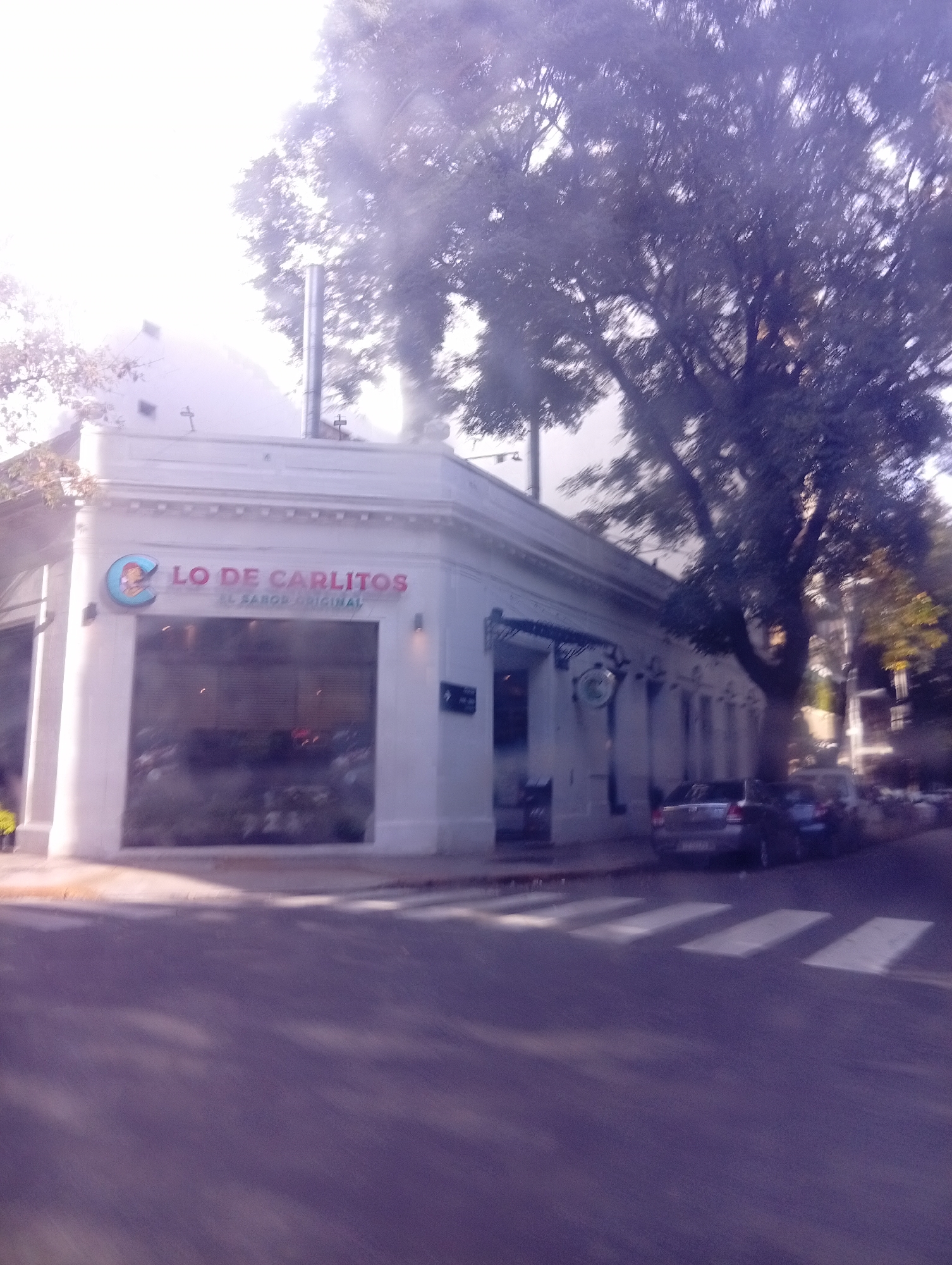 The image depicts the exterior of "Lo De Carlitos," a restaurant with a logo featuring a cartoon character. The building is a white corner structure with large windows and a sign that reads "Lo De Carlitos, El Sabor Original." Above the restaurant, a metal chimney can be seen.
Adjacent to the restaurant is a street with a pedestrian crossing marked by white stripes. Two cars, one grey and the other blue, are parked along the sidewalk. A large tree casts shadows on the street.
The image appears to be taken in daylight, with a slightly blurry or hazy quality, giving a sense of an urban environment. The style of the building and the name of the restaurant suggest that this is likely in Buenos Aires, Argentina.