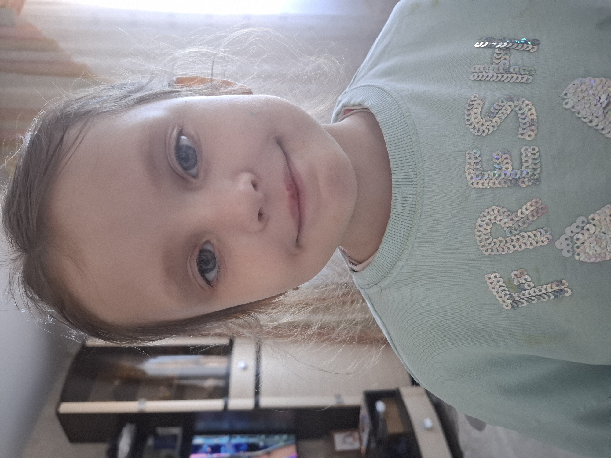 A young child is posing for a portrait indoors. The child has light skin, blue eyes, and light brown hair with some blonde highlights. She is wearing a light blue sweatshirt with the word "FRESH" written on it in silver sequins. The child has a slight smile.
The background features a cabinet with a dark brown base and beige upper section, suggesting an indoor setting in a residential area. While a precise location in Chaikovsky, Russia cannot be determined from the image alone, the interior design may be typical of homes in that region.