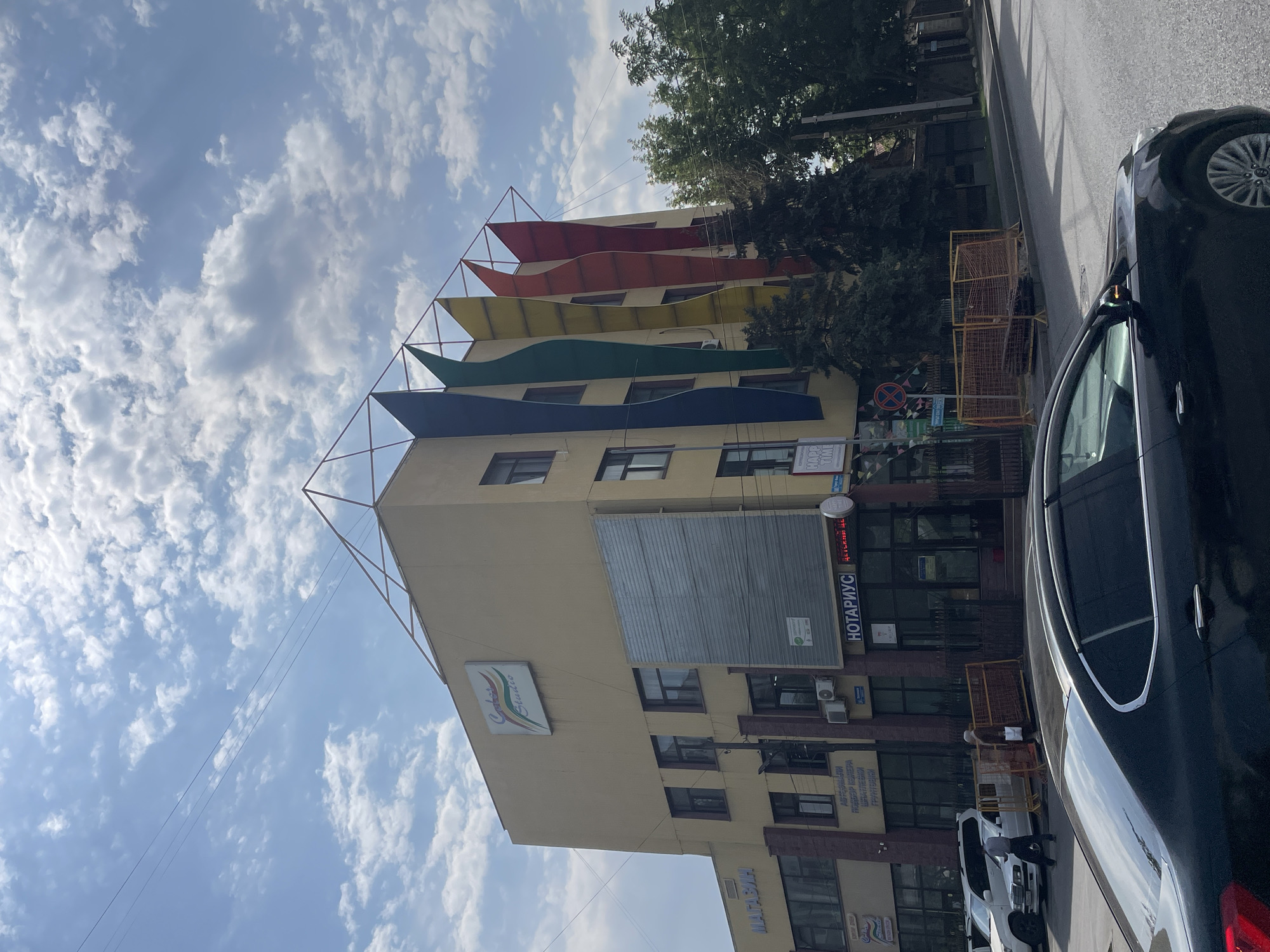 The image shows a view of a modern, multi-story building on a street in Almaty, Kazakhstan. The building has a light yellow facade and is decorated with large, colorful, wavy panels that resemble flames or feathers, in shades of blue, green, yellow, orange, and red, ascending from the roofline. A sign on the building reads "Color Studio". Below this, there is a large, metallic panel on the facade. Further down, on the ground floor, signage indicates a "Magazin" (Shop) and "Notarius" (Notary). A black car is parked in the foreground, with its rear partially visible. A white SUV and another white car are parked further down the street. Several people are walking on the sidewalk, and construction barriers are visible. Trees line the street and surround the building. The sky is blue with scattered white clouds.