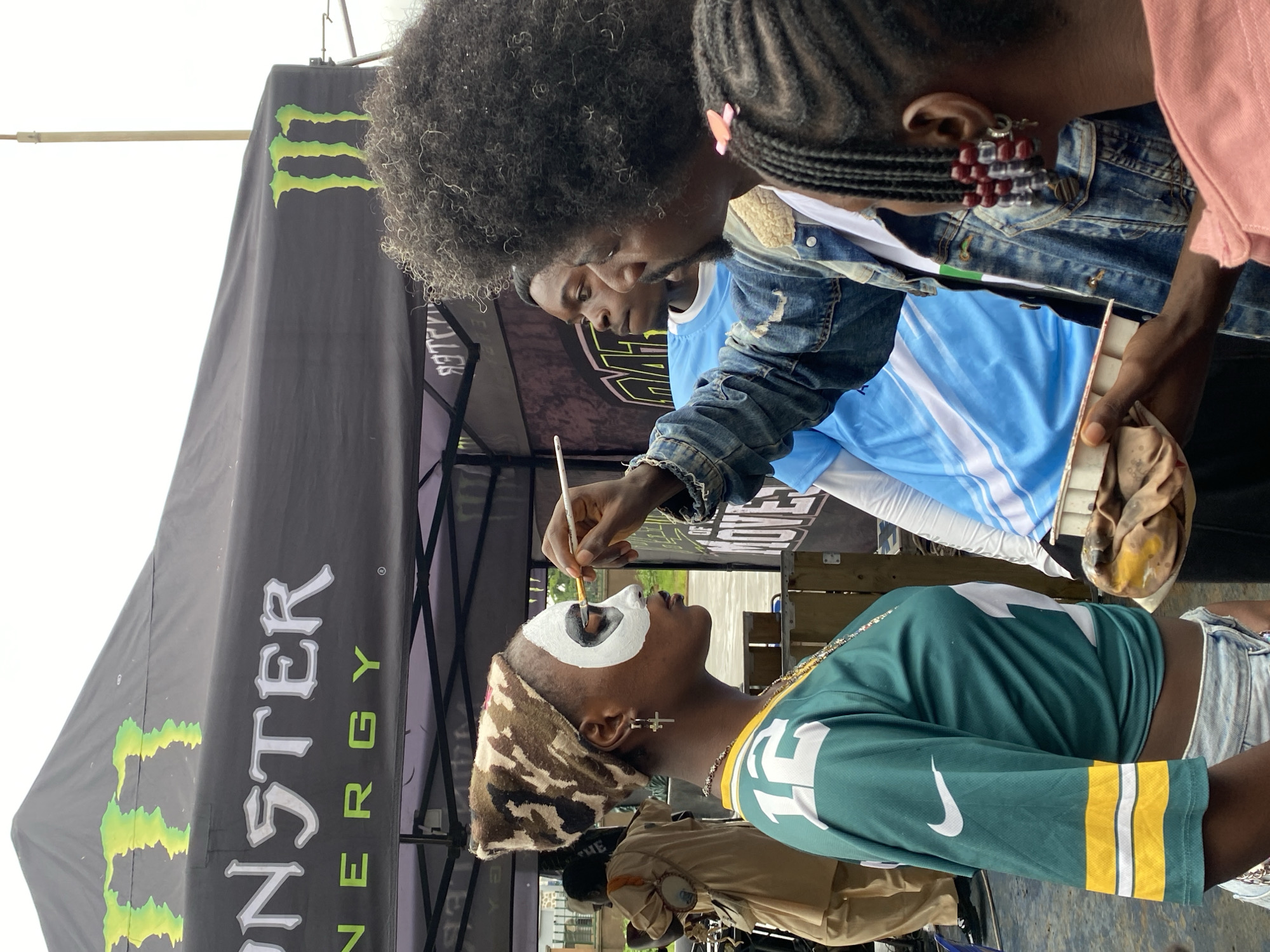 In the city of Gura Top, Nigeria, at an outdoor event likely associated with Monster Energy, a young woman is receiving face paint application. She is positioned in the foreground, facing left, with her head adorned in a camouflage-patterned bandana. Her face is partially painted with white and black makeup, suggesting a costume or artistic design. She wears a green and yellow jersey with the number "12" and a Nike logo.
To her right, a man with dark, curly hair is applying the face paint using a brush, holding a palette with makeup in his other hand. He is dressed in a denim jacket. Behind them, other individuals are visible, including a man in a blue shirt and another person in the background wearing a light brown outfit. A large black tent with the Monster Energy logo and branding is prominently displayed overhead, indicating the event's sponsor. The environment appears to be an open area, possibly a street or public space, with a hint of greenery and a cloudy sky.