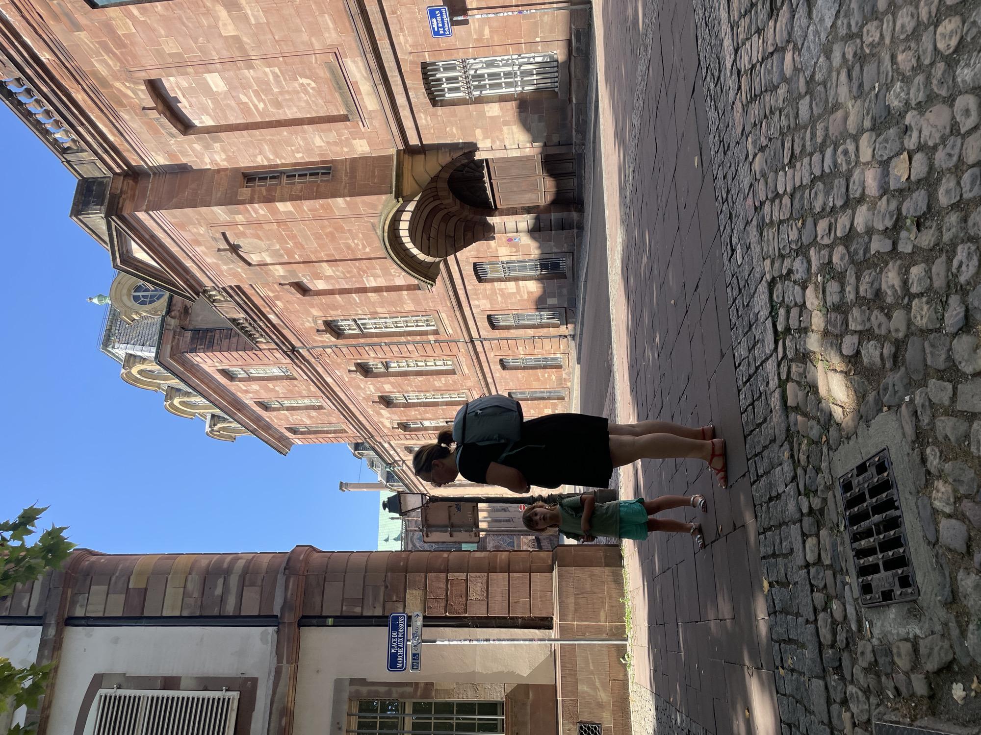 In Strasbourg, France, a woman wearing a black dress and a teal backpack stands with her back to the camera, looking towards the right. Beside her, a young child in a green shirt and turquoise shorts faces the camera, looking up with an open mouth. They are on a cobblestone street in front of a large, ornate building constructed of red stone. A blue street sign with white lettering reads "PLACE DU MARCHE AUX POISSONS" and an arrow pointing to the right. The building features arched doorways, barred windows, and decorative architectural elements. The sky above is clear and blue.