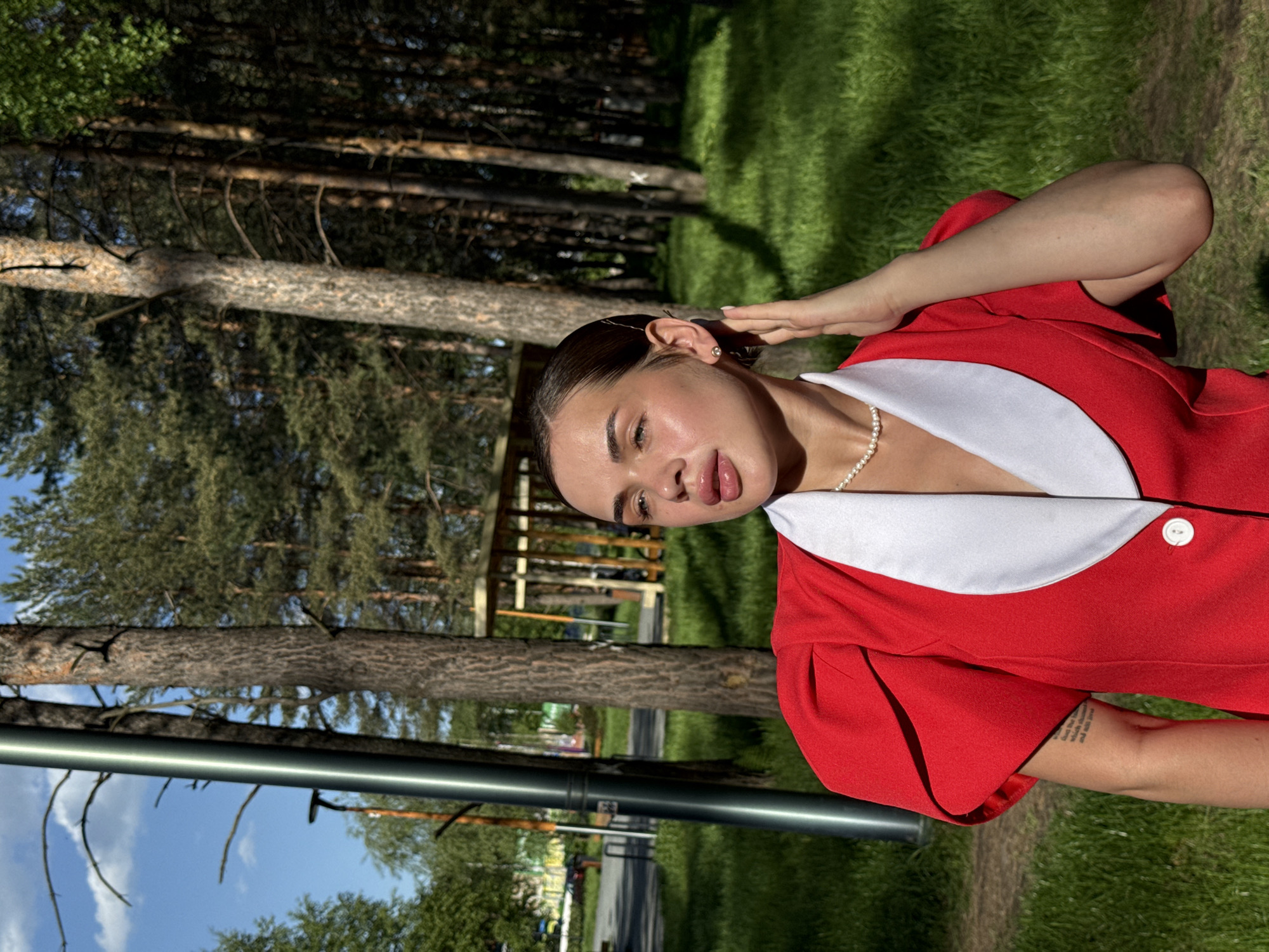 A medium outdoor shot shows a woman in Perm, Russia. She wears a red jacket with a white collar, a pearl necklace, and an earring in her left ear. Her left hand is raised behind her head. Trees and greenery are visible in the background, with a building in the distance. A gray metallic bar is partially visible in the lower left of the frame. The sun is out, as there are cast shadows.