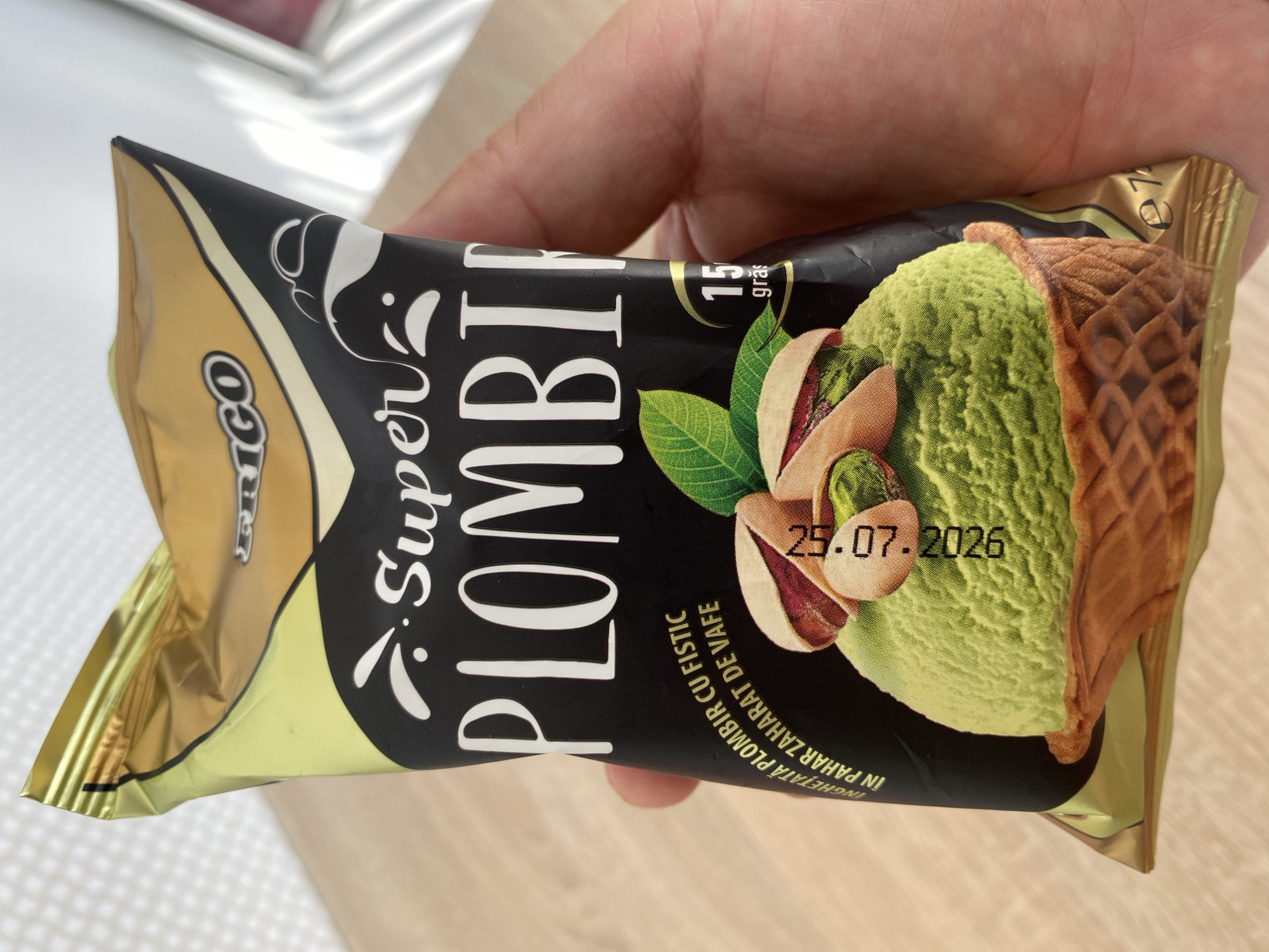 A person is holding a package of "Super Plombir" pistachio ice cream. The package has a black background with white lettering and images of pistachios and a green ice cream cone. The text on the package is in Romanian and indicates that it is pistachio ice cream in a waffle cone. There is also a date printed on the package: 25.07.2026. The background is a wooden table. There is no visible information in the image that would allow for the location to be determined as Chișinău, Moldova.