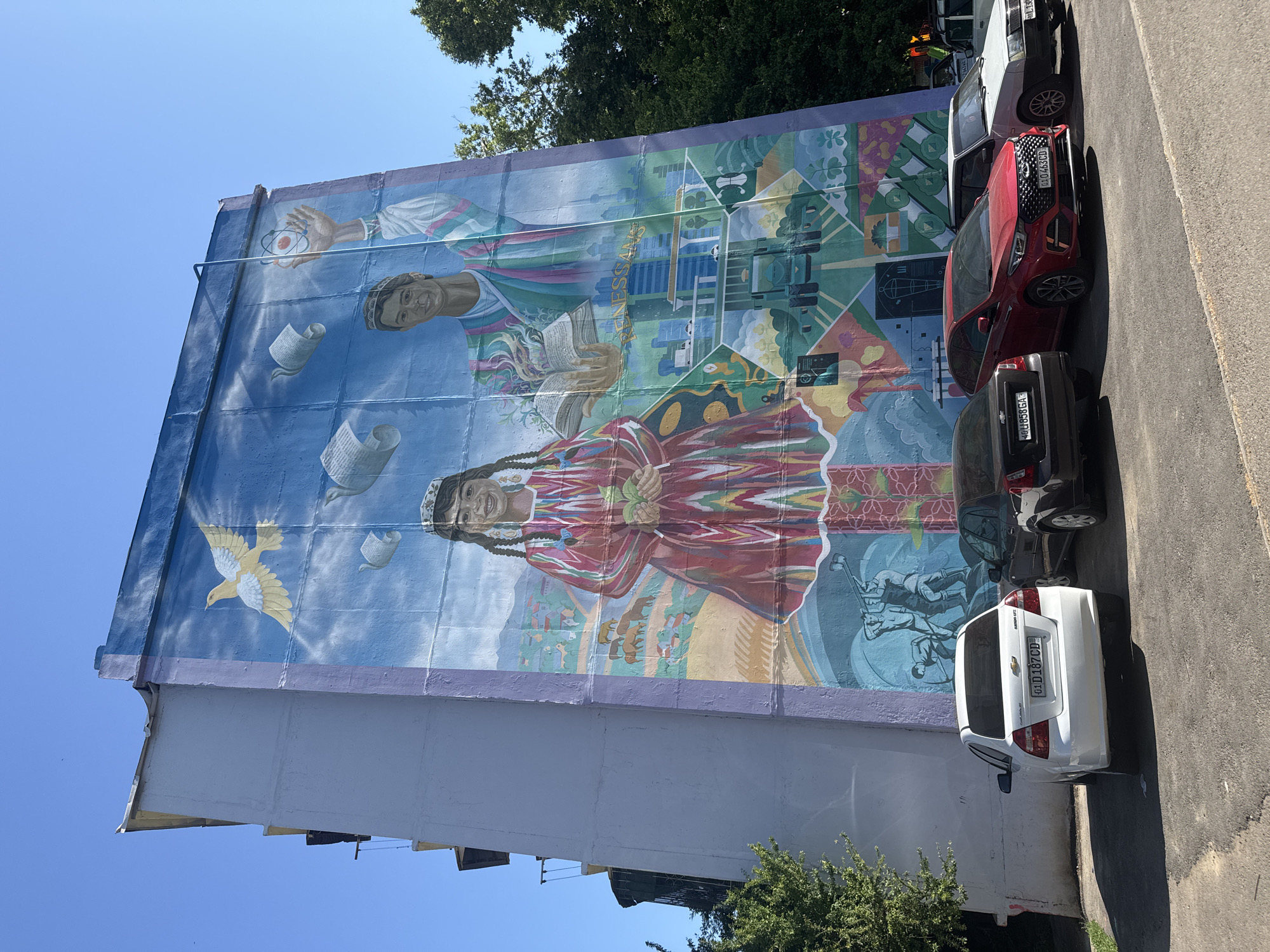 A large mural adorns the side of a building in Tashkent, Uzbekistan. The mural features two prominent figures: a woman and a man, both appearing in traditional Uzbek attire. The woman is depicted holding fruits, wearing a colorful robe and traditional headdress. The man is portrayed holding a book and an atom model, also wearing traditional clothing. The background of the mural includes images of modern architecture, solar panels, and other symbols.
The mural is painted on the side of a multi-story building with a light-colored facade. Several cars are parked along the street adjacent to the mural, including a white Chevrolet, a dark gray car, and a red Kia. The street is paved, and some greenery is visible at the base of the building and overhead. The sky is clear and blue.