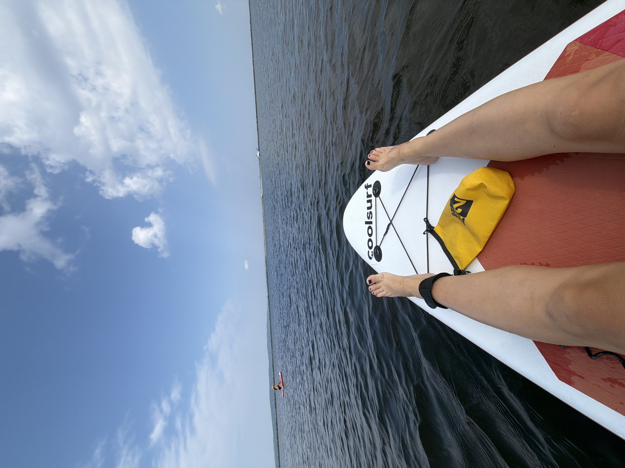 A first-person perspective shot shows a person's legs and feet resting on a white paddleboard in the water. The paddleboard has the logo "coolsurf" on it. A yellow dry bag is placed on the board near the person's feet. The water is dark and choppy, with a horizon line in the distance. The sky is blue with scattered white clouds. Another person is visible on a paddleboard further in the distance. The location is the Kama River in Chaikovsky, Russia, as indicated by the presence of the Kama River and the city's geographical context.