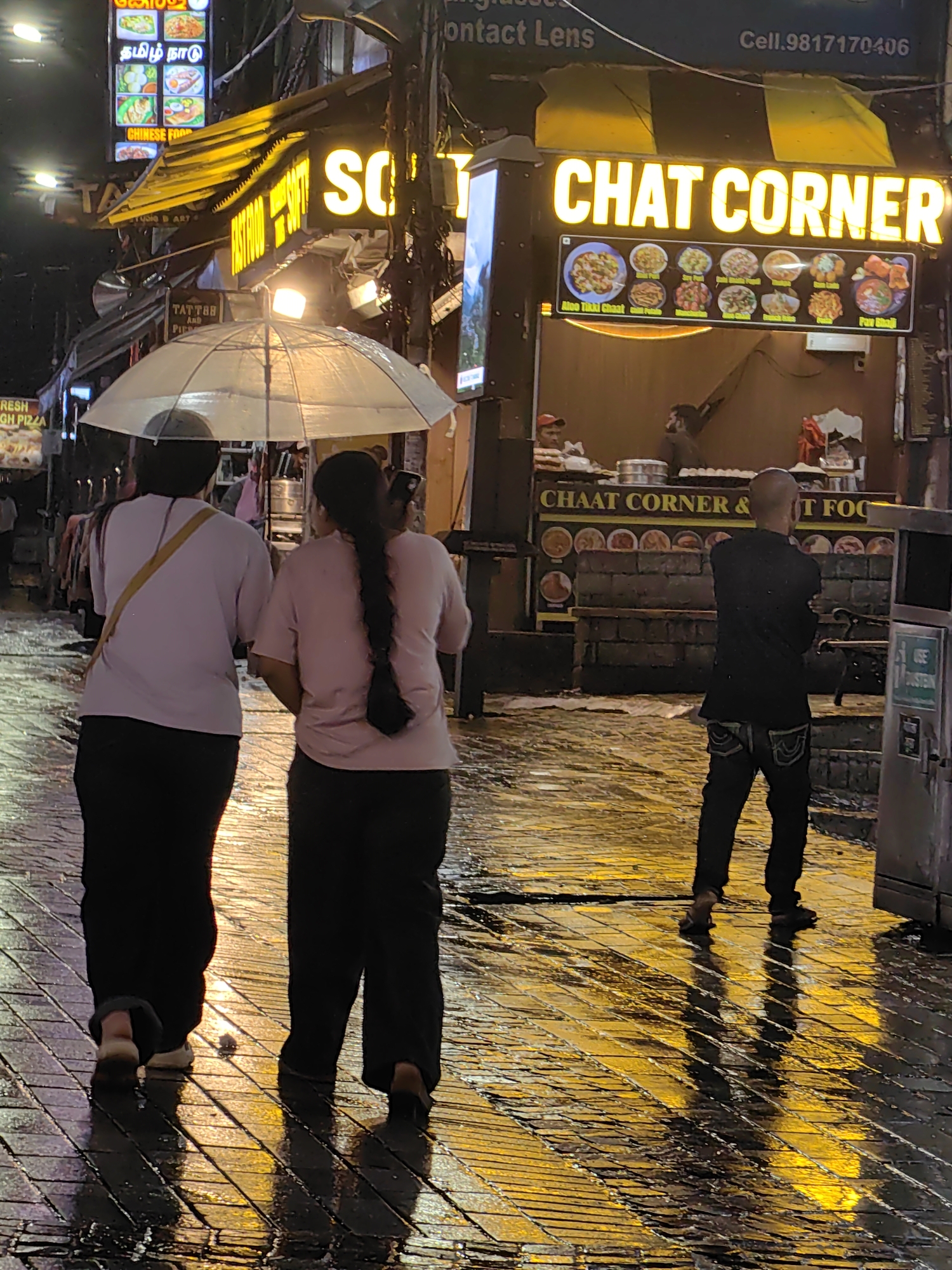 The image depicts a street scene in Manali, India, at night. The ground is wet, reflecting the bright yellow neon lights of a "CHAT CORNER" establishment. Two individuals, viewed from behind, walk under a clear umbrella. To their right, a bald man stands with his arms crossed, also facing away from the camera. The "CHAT CORNER" sign is illuminated, displaying various food items. Further to the left, another sign advertises "CHINESE FOOD" with an array of dishes shown. The overall atmosphere suggests a bustling street market or food stall area, potentially experiencing rain.