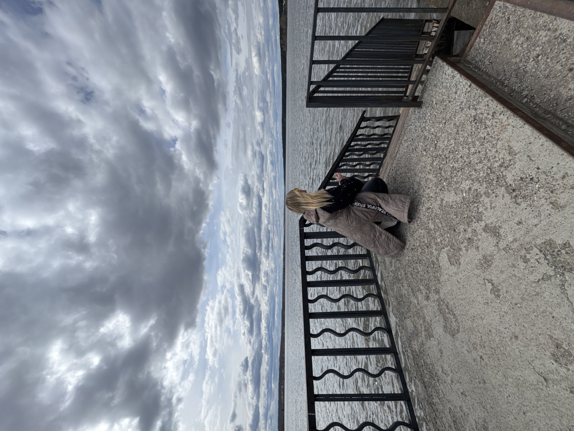 The image, taken in Votkinsk, Russia, captures a person sitting on a concrete pier or embankment overlooking a vast body of water under an overcast sky. The individual, who appears to be a woman with long blonde hair, is seen from behind, gazing out towards the horizon. She is wearing a dark top and light-colored trousers or a long jacket. Part of the text "BEAUTIFUL STUDI" is visible on her clothing.
Along the edge of the concrete structure, to the person's right, a black metal railing with a distinctive wavy design runs parallel to the water. Further to the right and slightly set back, another black metal railing, featuring a more angular and grid-like pattern, suggests a pathway or stairs leading to a lower level or further along the pier.
The extensive body of water, likely a lake or wide river, stretches into the distance, meeting a faint, flat shoreline on the opposite side. The surface of the water shows ripples, indicating slight movement or wind. Above, the sky is largely covered by grey, textured clouds, though several patches of lighter blue sky are visible through breaks in the cloud cover, suggesting it is daytime. The overall scene evokes a moment of quiet observation or contemplation by the water.