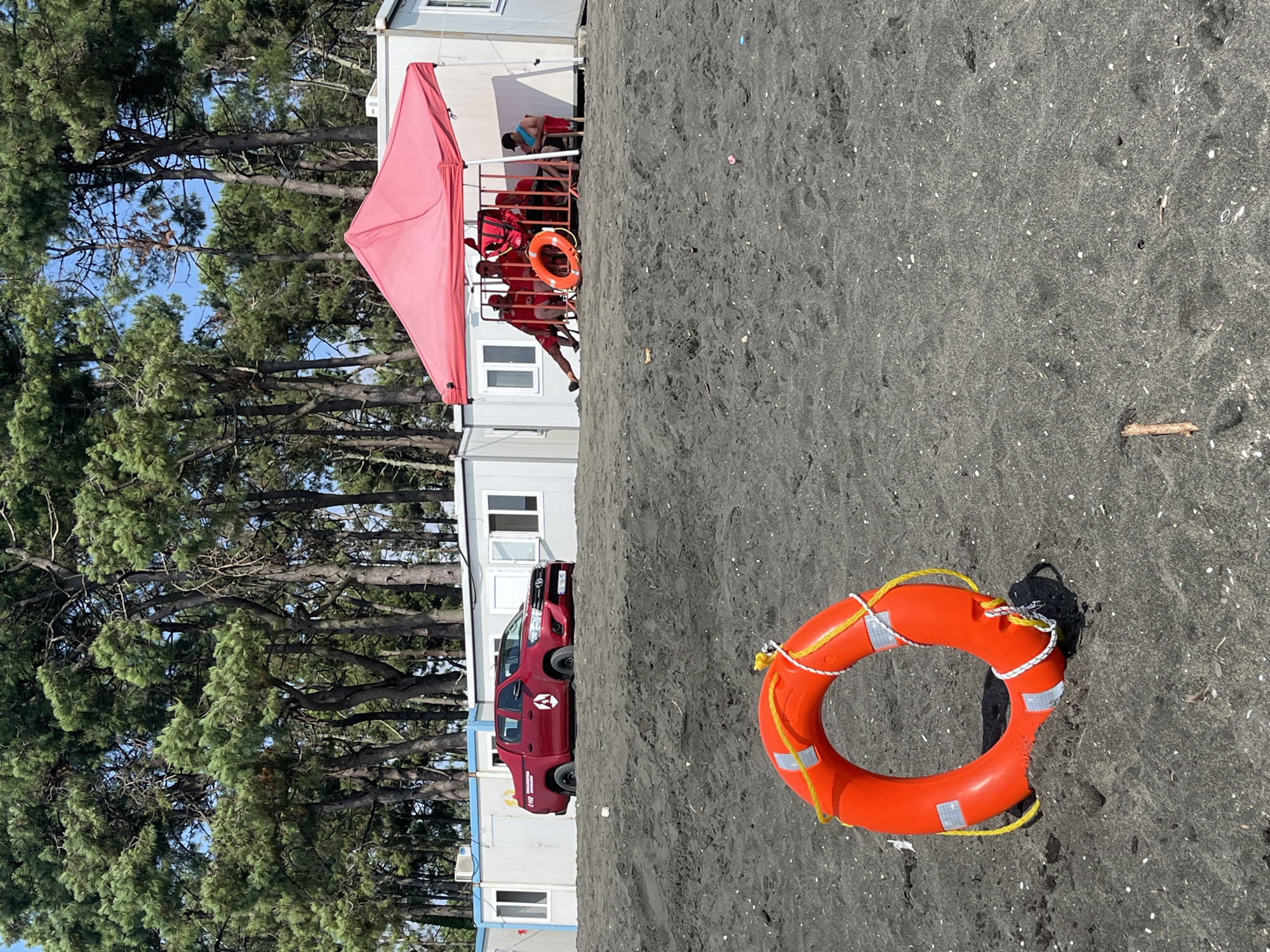 This image captures a scene on the black sand beach of Ureki, Georgia. In the foreground, a bright orange life preserver with yellow rope and reflective tape is partially embedded in the sand. In the middle ground, a red pickup truck with "0112" and a white triangular logo on its side is parked on a sandy incline. Behind the truck, a row of modular white buildings with windows and doors lines the edge of the beach. A red canopy is erected in front of one of these buildings, providing shade for several individuals wearing red shirts, some of whom appear to be lifeguards. One person sits alone on a chair to the right of the canopy. The background is dominated by a dense forest of tall, green pine trees. The sky above is clear and blue.