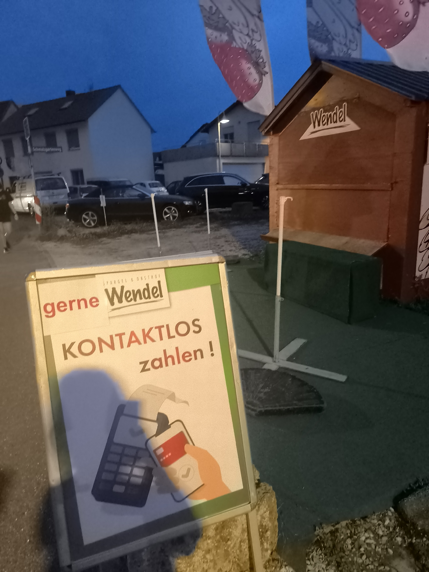 The image depicts an outdoor scene at dusk, featuring a sign in the foreground. The sign is white with green and orange text. The text on the sign reads "gerne Wendel" in orange and black, followed by "KONTAKTLOS zahlen!" in large orange letters and "zahlen!" in smaller orange text. Below the text, there is an illustration of a mobile phone and a card reader, with a credit card being inserted into the phone.
Behind the sign, a wooden structure resembling a small stand or booth is visible, with the word "Wendel" painted on it in white lettering within a triangle. The structure appears to be made of dark wood. A white pole with a hook is planted in front of the booth.
In the background, houses and parked cars are visible. The sky is dark blue, indicating evening or twilight. There are also large banners with strawberry illustrations hanging from unseen poles, adding a decorative element to the scene. The ground appears to be a mix of asphalt and gravel.