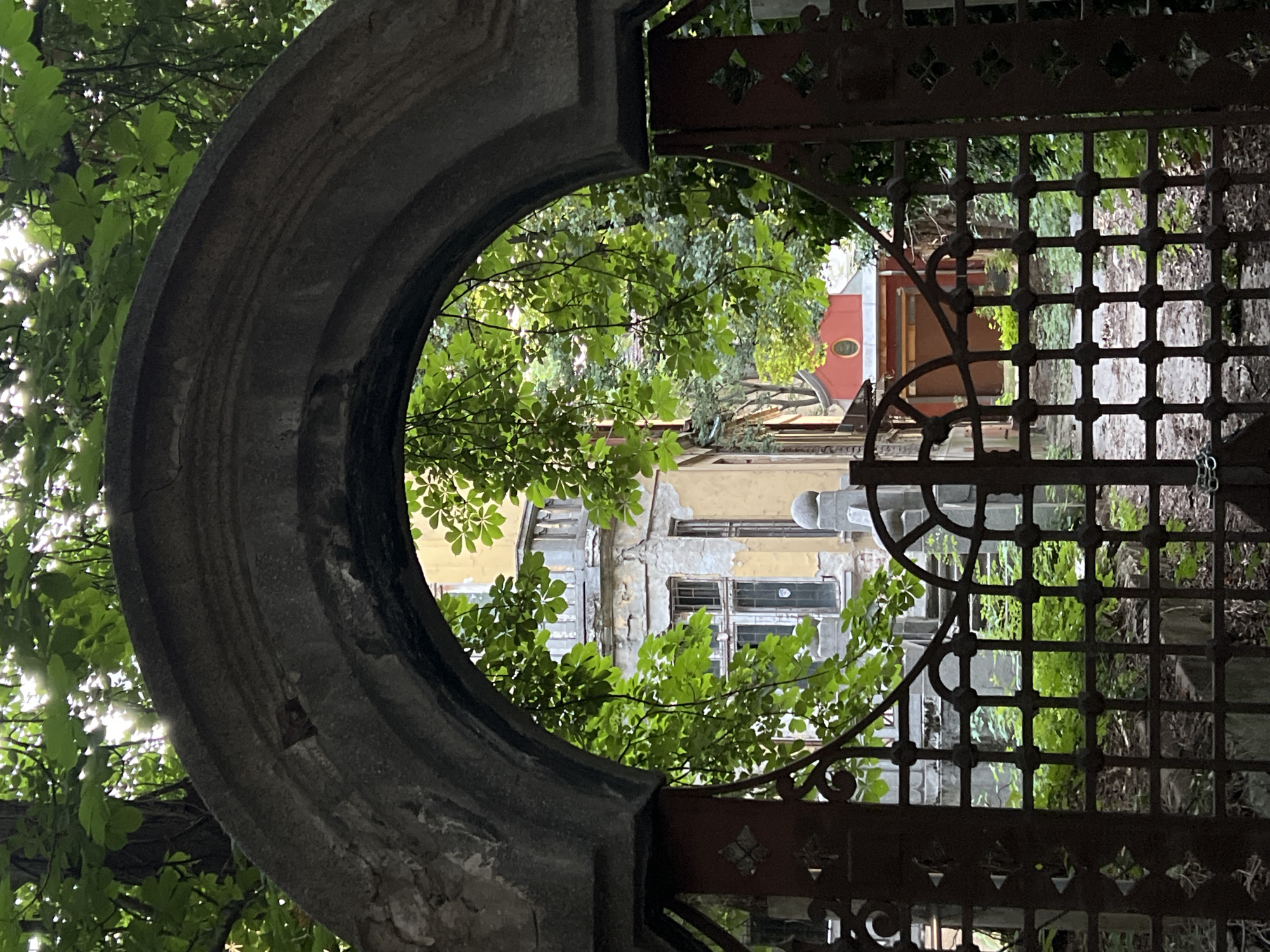 The image captures a view through a weathered stone archway and an ornate, rusty metal gate, revealing a glimpse of an old courtyard in Sofia, Bulgaria. The archway, constructed of rough, grey stone, is partially obscured by lush green foliage from overhanging trees, with sunlight filtering through the leaves.
The gate in the foreground is made of dark, wrought iron with intricate geometric patterns and a central decorative element. Beyond the gate, the courtyard is overgrown with greenery. In the background, an aged, pale yellow building with balconies and shuttered windows stands, showing signs of decay with peeling paint and crumbling plaster. To the right of this building, a smaller structure with a red roof and an oval window is visible. The ground within the courtyard appears to be paved or covered in gravel and fallen leaves, with more greenery emerging from the cracks. The overall impression is one of neglect and the passage of time, with nature reclaiming a historically significant space.