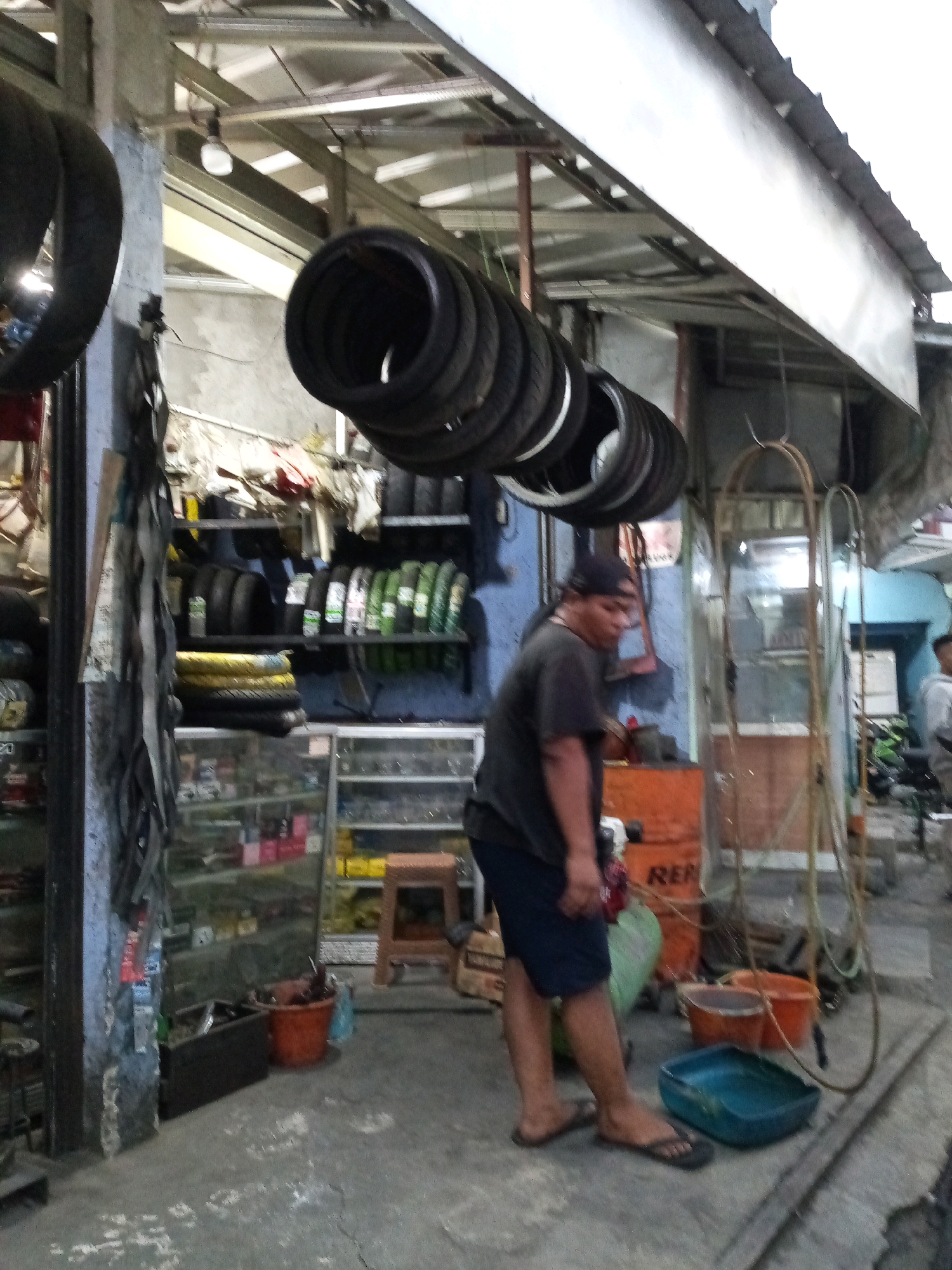 This is a medium-shot, slightly low-angle, full-color photograph of a roadside tire shop in Bekasi, Indonesia. The scene is cluttered with tires and various shop items. In the foreground, a person wearing a black t-shirt, dark shorts, and flip-flops stands facing left, looking down. They are near a bright orange bin. Behind this person, to the right, are two orange buckets and a blue plastic container on the ground.
The background reveals shelves filled with tires and other products. Tires are also hanging from the ceiling structure above the shop. The shop appears to be an open-air establishment, with a corrugated metal roof supported by metal beams. The ground is concrete. The lighting suggests it is daytime, possibly overcast or late afternoon. The overall impression is of a busy, functional workshop.