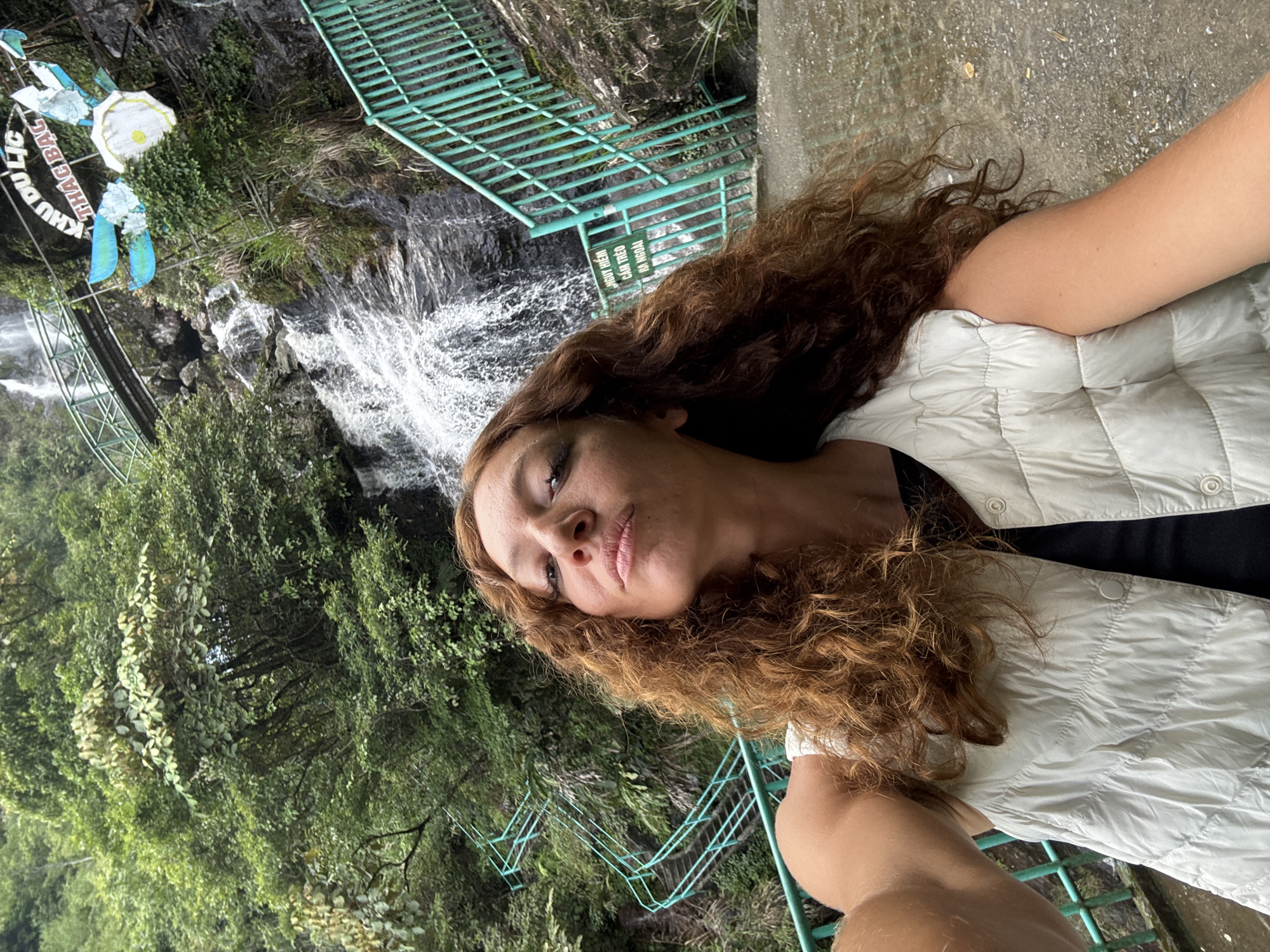A selfie-style, medium shot captures a woman with curly brown hair, wearing a white quilted vest over a black top, positioned at an angle in front of a waterfall and forested area. A portion of the concrete walkway is visible at the top right corner. A cascading waterfall flows over rocks, flanked by lush green trees. Teal metal stairs are seen at the top-left and bottom-middle of the frame. A sign with the words "KHU DU LICH THAC BAC" is present on the upper-left, suggesting this is the Silver Waterfall tourist attraction in Sa Pa Ward, Vietnam.