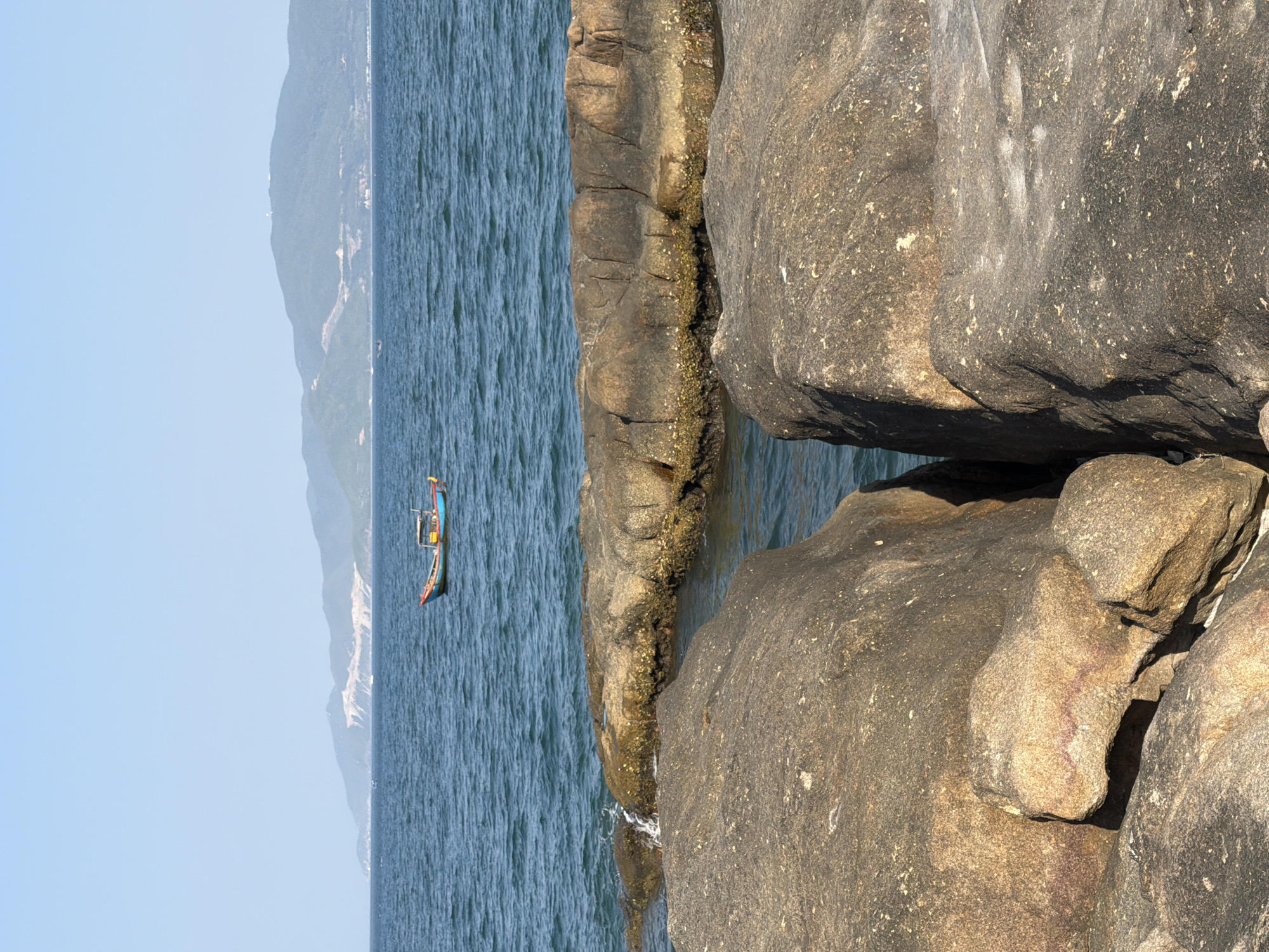 The image displays a coastal scene in Nha Trang, Vietnam, under a clear blue sky. In the foreground, large, rough-textured, brown and grey rocks dominate the view, with a narrow channel of calm blue water visible between two prominent rock formations. The water is clear enough to reveal submerged rocks or sand. Further out, the ocean stretches towards a hazy mountainous coastline. A small, colorful fishing boat with a blue hull and yellow and orange accents is anchored in the middle ground, bobbing gently on the rippling blue water. The distant mountains are covered in vegetation, and signs of human activity, possibly construction or roads, are visible on their slopes. The overall impression is one of natural beauty and a peaceful coastal environment.