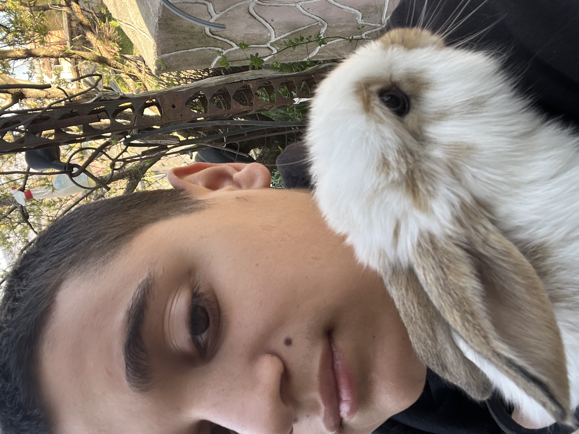 A close-up, slightly angled shot features a person and a rabbit. The person, likely male, is fair-skinned with short, dark hair and distinct eyebrows. Their right eye is visible and their expression is neutral. A mole is noticeable on the left side of their nose. The rabbit, predominantly white with brown markings around the ears and eyes, is positioned close to the person, possibly being held. The background is somewhat blurred, showing elements like a weathered metal structure, vegetation, and a paved surface. Based on the location provided, this scene is situated in Lozarevo, Bulgaria. There are no further details available to describe the specifics of the setting.