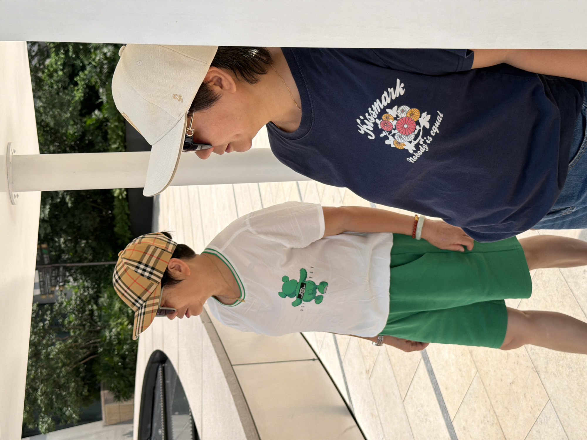 Two people stand outdoors in Yuzhong District, China. The person on the left wears a white t-shirt with a green teddy bear graphic, green shorts, and a tan, brown, and black plaid baseball cap. The person on the right wears a navy blue t-shirt with a floral graphic and text that reads "Kissmark nobody is equal," and a light beige baseball cap. Both individuals wear sunglasses. The ground is paved with light-colored square tiles, and a white pillar is visible between the two individuals. In the background, trees and modern architecture are present.
