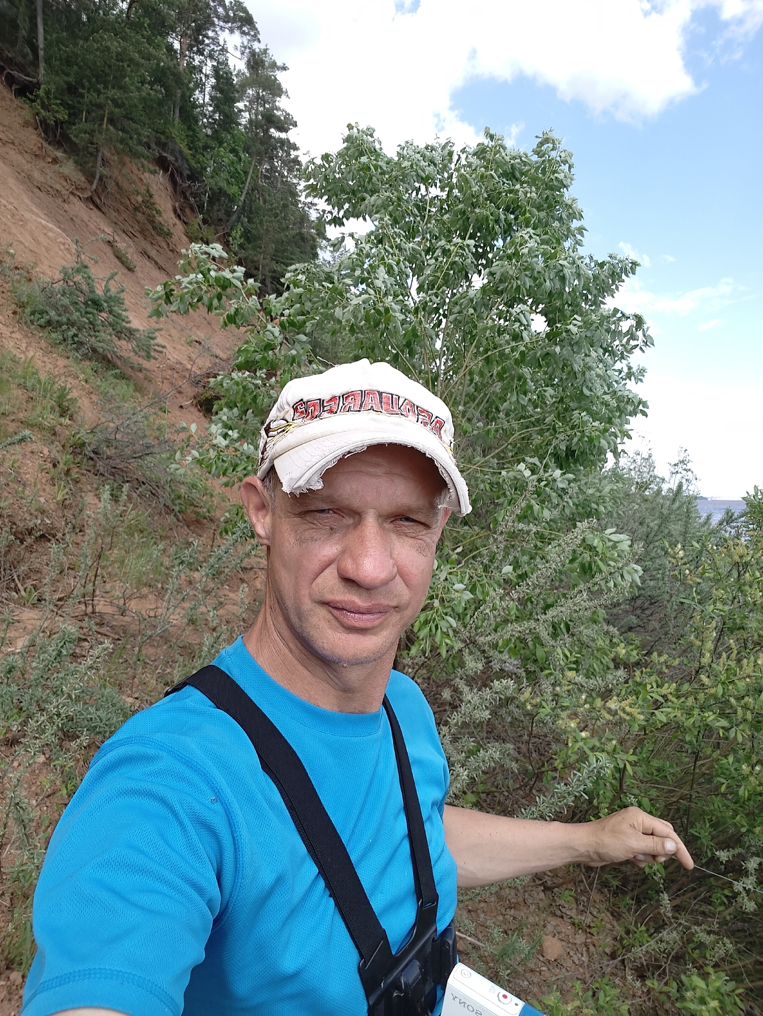 A man in a blue t-shirt and a worn, white baseball cap stands in a natural setting. The background features a hillside covered with trees and vegetation on the left, and a tree with dense foliage on the right. In the distance, a glimpse of a body of water under a sky with scattered clouds can be seen. The man is pointing with his right hand towards the vegetation on the right. He has a camera slung across his chest. The location is in the city of Прикамский, Russia.