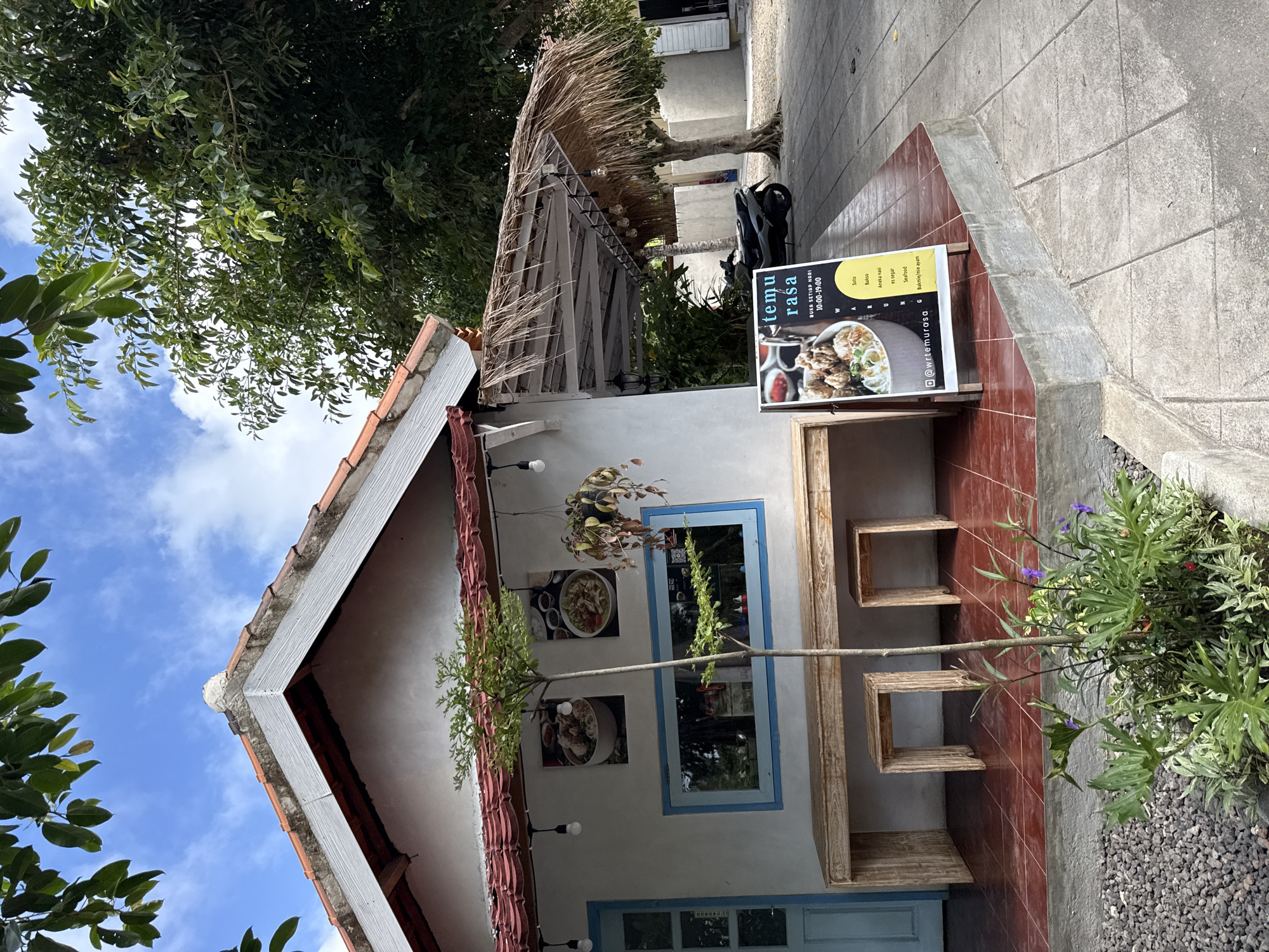 Here's a description of the image:
The image depicts the exterior of a building in Pejukutan, Indonesia. The building has a white facade with blue window and door frames, and a tiled roof. A menu board with the words "Temu Rasa" is visible next to the building, advertising various food items such as Soto, Bakso, and Seafood. A wooden shelf-like structure with cube cutouts is attached to the wall. Greenery is present around the building and in hanging planters. The sky is visible in the background.