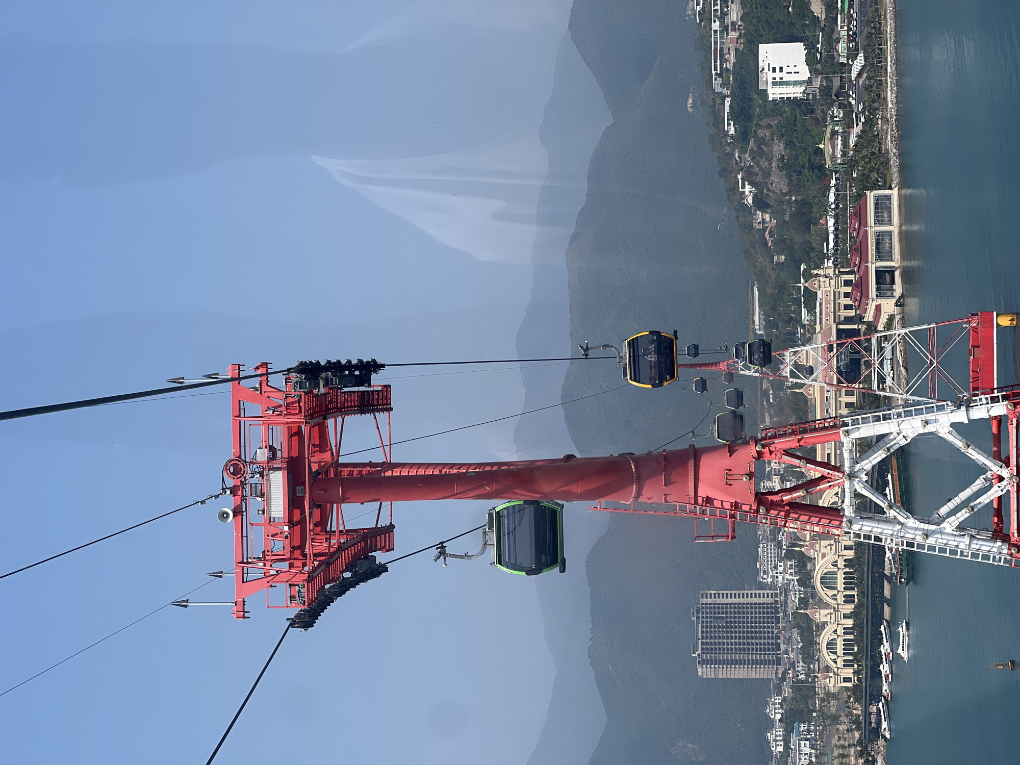 The image depicts a cable car system, likely in Nha Trang, Vietnam, based on the coastal city backdrop. A red and white metal tower anchors the cables, with several gondolas suspended along the line. The gondolas are varying colors. The background features mountains, a coastline with a city, and the sea. The sky is clear.