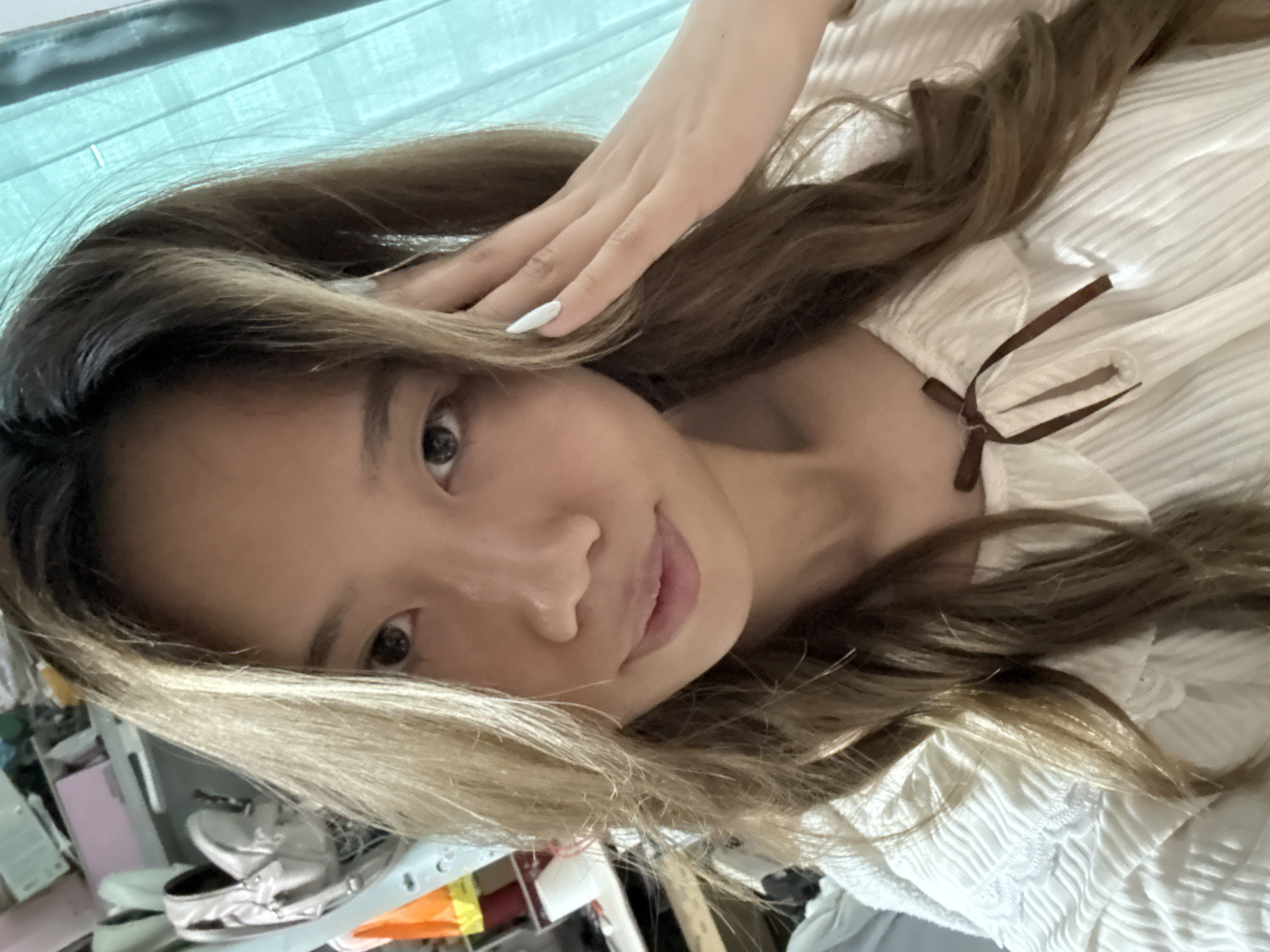 A young woman poses for a selfie. She is fair-skinned with brown hair and light blonde highlights. Her right hand rests on her head. She is wearing a textured white top with a brown ribbon detail. The background includes a teal curtain and a cluttered desk. It is impossible to identify the exact location in Nan'an District from the visual information provided.