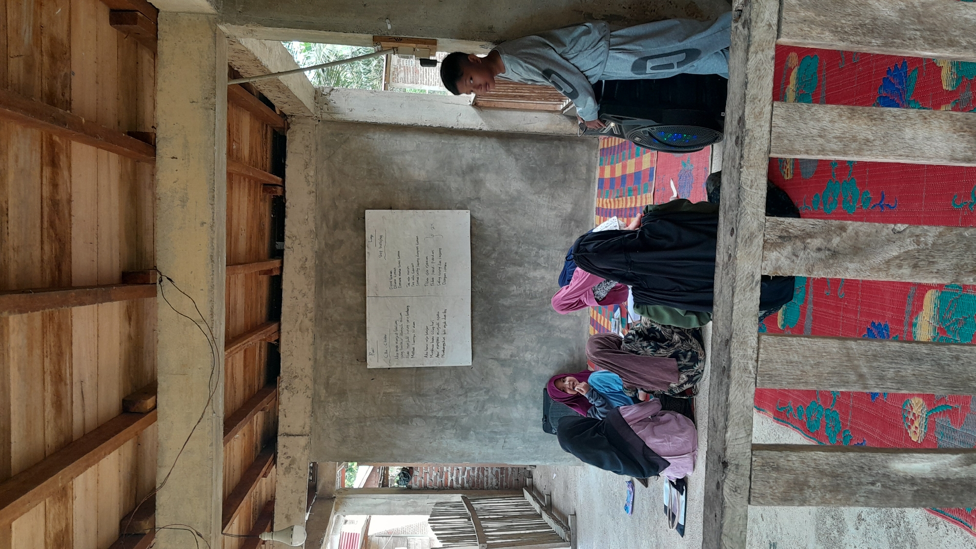 The image captures an indoor scene, likely a classroom or meeting room, in the city of Aluerambee, Indonesia. A young boy stands near a speaker, seemingly adjusting it. Several females, wearing traditional Muslim clothing like hijabs, are seated on the floor. On the wall, there is a whiteboard containing hand-written notes or lessons in the local language. The room's structure features a mix of concrete and wooden elements. The presence of woven mats on the floor suggests it is a communal space.