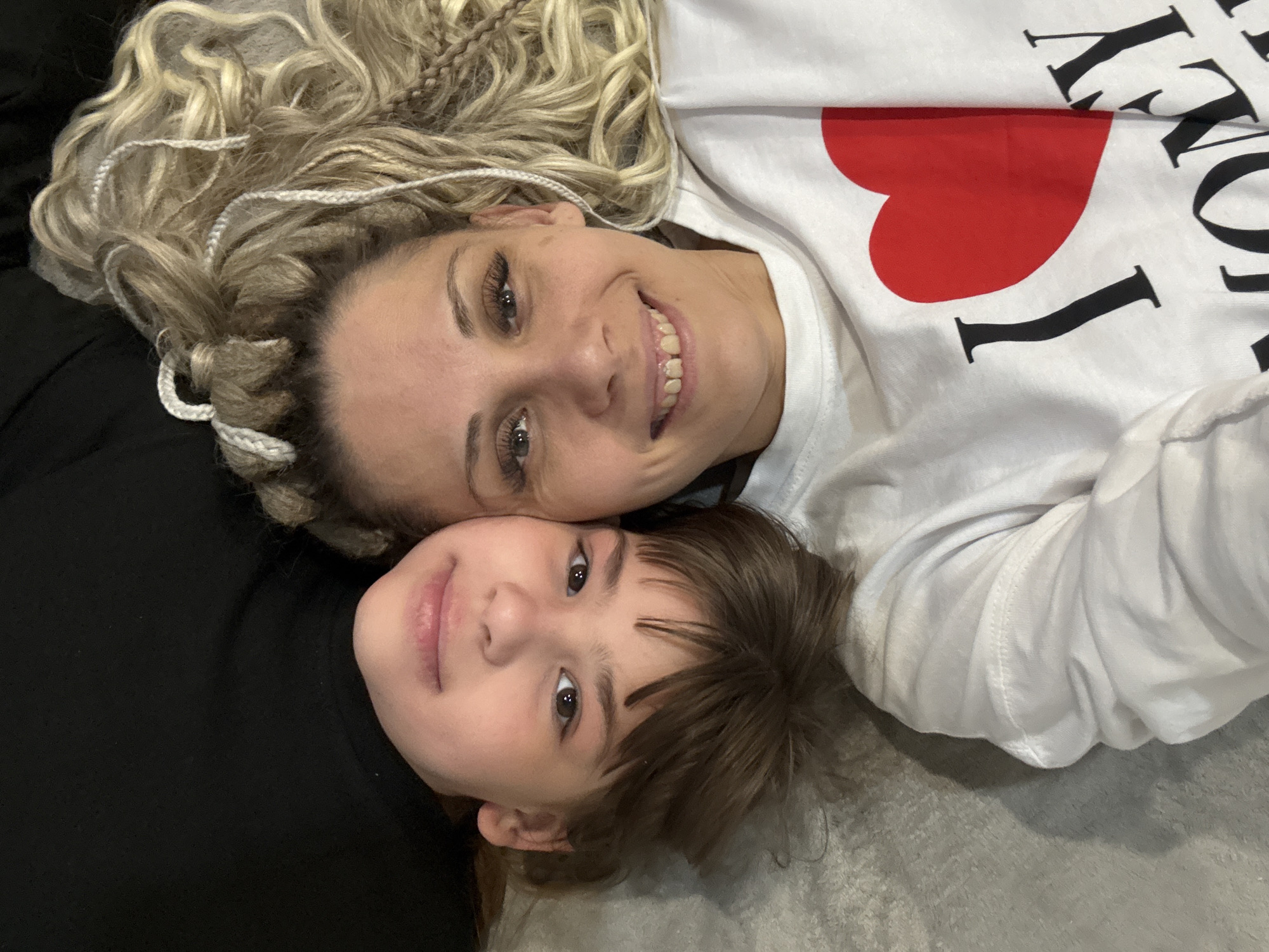 A selfie depicts an adult female and a child lying down. The woman has blonde hair with white braids, is wearing a white top with a red heart and the words "I Love ROCKY," and is smiling. The child has brown hair and is wearing a black top. The background is a grey surface. The location is unidentified with no evidence to indicate it is Rusanovo, Russia.