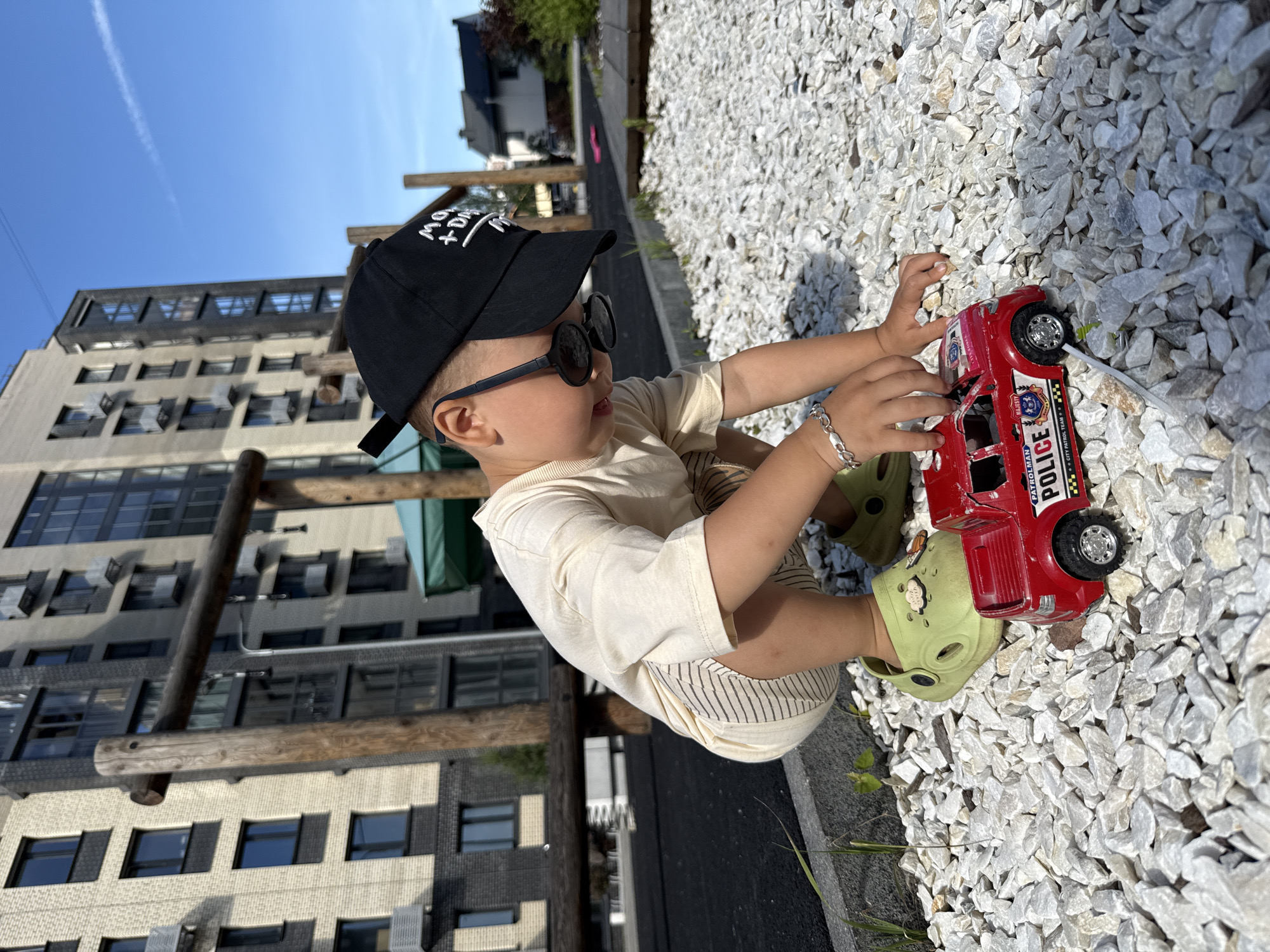 A young boy, wearing a light-colored t-shirt, striped shorts, and lime green crocs, crouches down on a playground covered in white gravel. He is wearing a black baseball cap and sunglasses pushed up on his forehead. The boy is holding a red toy police car, its siren light clearly visible. In the background, a modern, multi-story apartment building with dark-framed windows stands under a clear blue sky. Wooden poles, part of a playground structure, are in the foreground. The scene is illuminated by bright sunlight, casting shadows on the gravel. The location is identified as the city of Rusanovo, Russia.