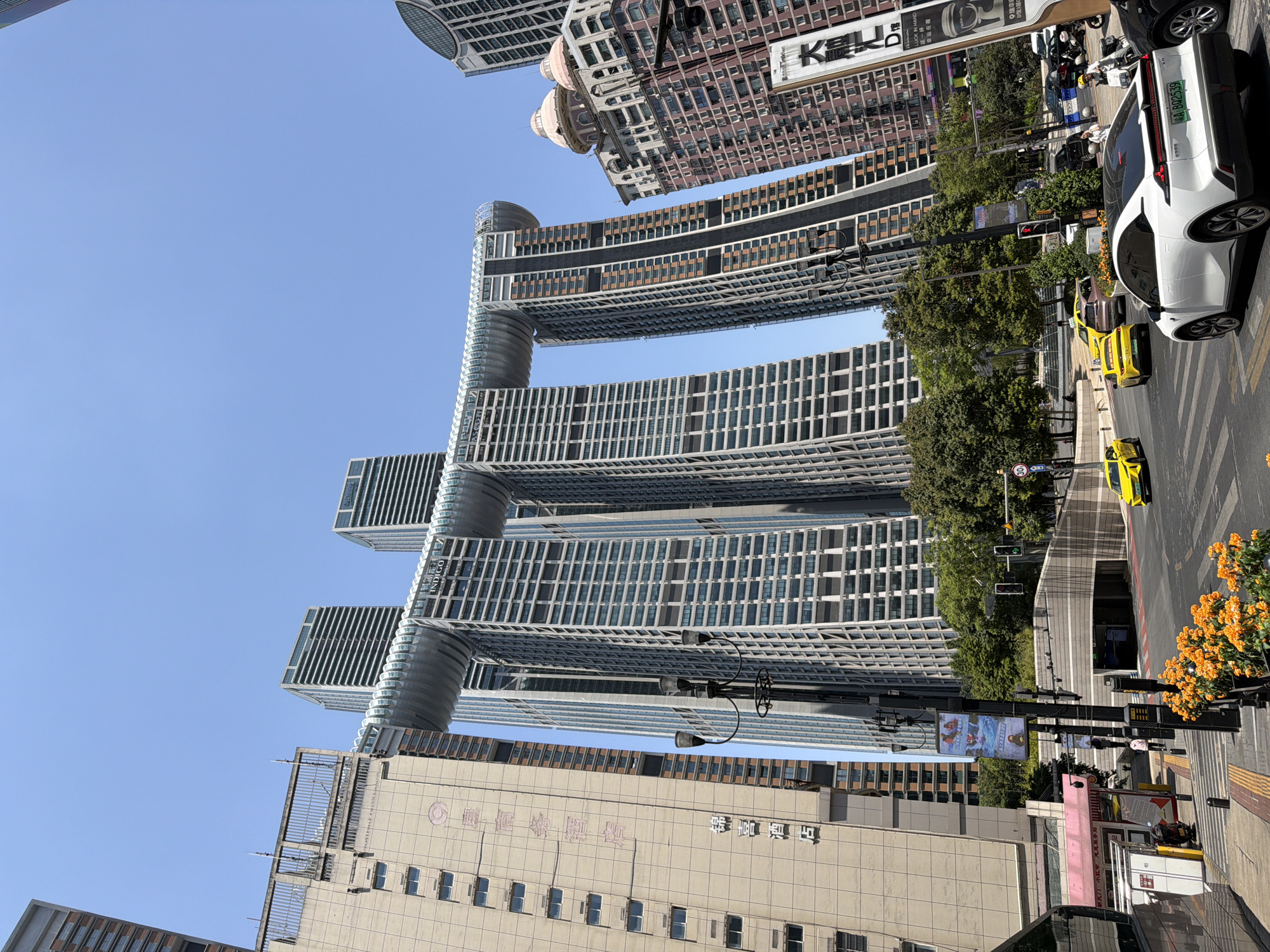 The image depicts a street scene in Yuzhong District, China, with a focus on the distinctive Raffles City Chongqing complex. The complex features two massive skyscrapers connected at their summits by a horizontal structure that resembles a ship or a dragon. The architecture is modern and imposing, with numerous windows reflecting the bright, clear blue sky.
In the foreground, a multi-lane road is visible with several vehicles, including a white electric car with a license plate that reads "A·B02539", two yellow taxis, and other cars. Pedestrians are present, some walking on the sidewalk and others near a crosswalk. A street light with multiple lamps stands prominently.
To the left of the Raffles City complex, a beige building with visible Chinese characters, possibly a hotel, is present. Further to the right, other buildings with diverse architectural styles, including some with pink domed roofs, are also in view. Lush green trees line parts of the street, contrasting with the urban landscape. The overall scene suggests a busy urban environment during daylight hours.