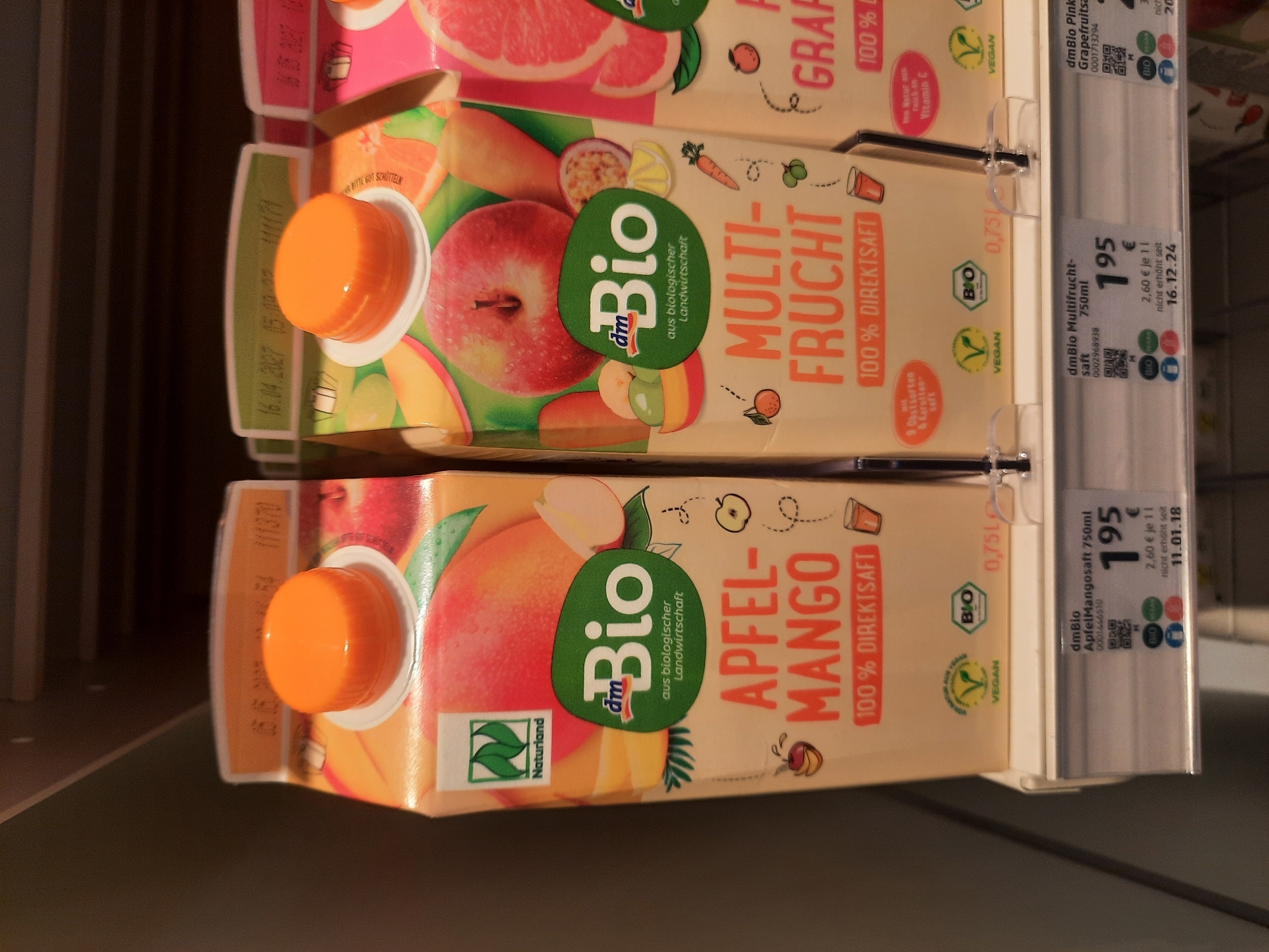 The image shows a shelf in a German supermarket with two cartons of dmBio brand juice. The carton on the left is labeled "Apfel-Mango 100% Direktsaft" (Apple-Mango 100% Direct Juice) and the carton on the right is labeled "Multi-Frucht 100% Direktsaft" (Multi-Fruit 100% Direct Juice). Both cartons are orange and feature images of the fruits they contain. Below the juice cartons, price labels are visible, with the dmBio ApfelMango Saft 750ml priced at 1.95 €. The background suggests a retail environment, possibly a dm-drogerie markt based on the branding. The scene is well-lit, indicating an indoor setting. No people are visible in the image. The time stamp on the price label for the ApfelMango Saft reads 11.01.18, suggesting this photograph was taken or uploaded on or after January 11, 2018. The location in Rosenheim, Germany is not depicted in the image itself, but is a contextual detail provided with the request.