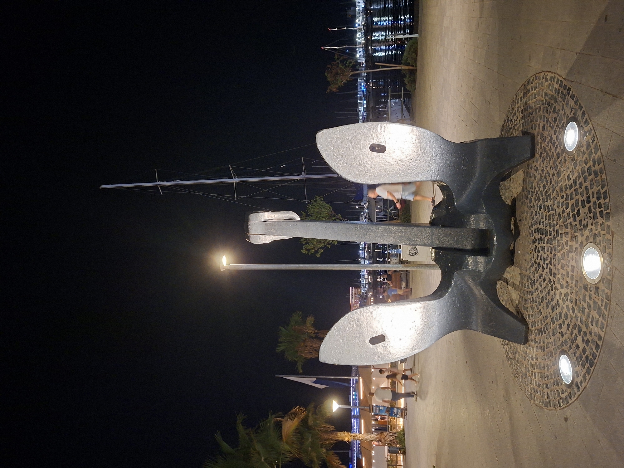 This is a nighttime photograph of a large, dark gray anchor sculpture located on a paved promenade in Alicante, Spain. The anchor is positioned in the foreground, centered in the frame, and illuminated by spotlights embedded in the circular mosaic base surrounding it. Behind the anchor, a tall sailboat mast pierces the dark night sky. To the left, palm trees and building lights suggest a coastal urban environment. Several people are visible in the background, walking along the promenade or seated at tables, indicating a public space. The water of a harbor is visible to the right, reflecting the lights of buildings and boats. The overall scene depicts a marina or waterfront area at night, with the anchor sculpture serving as a prominent focal point.