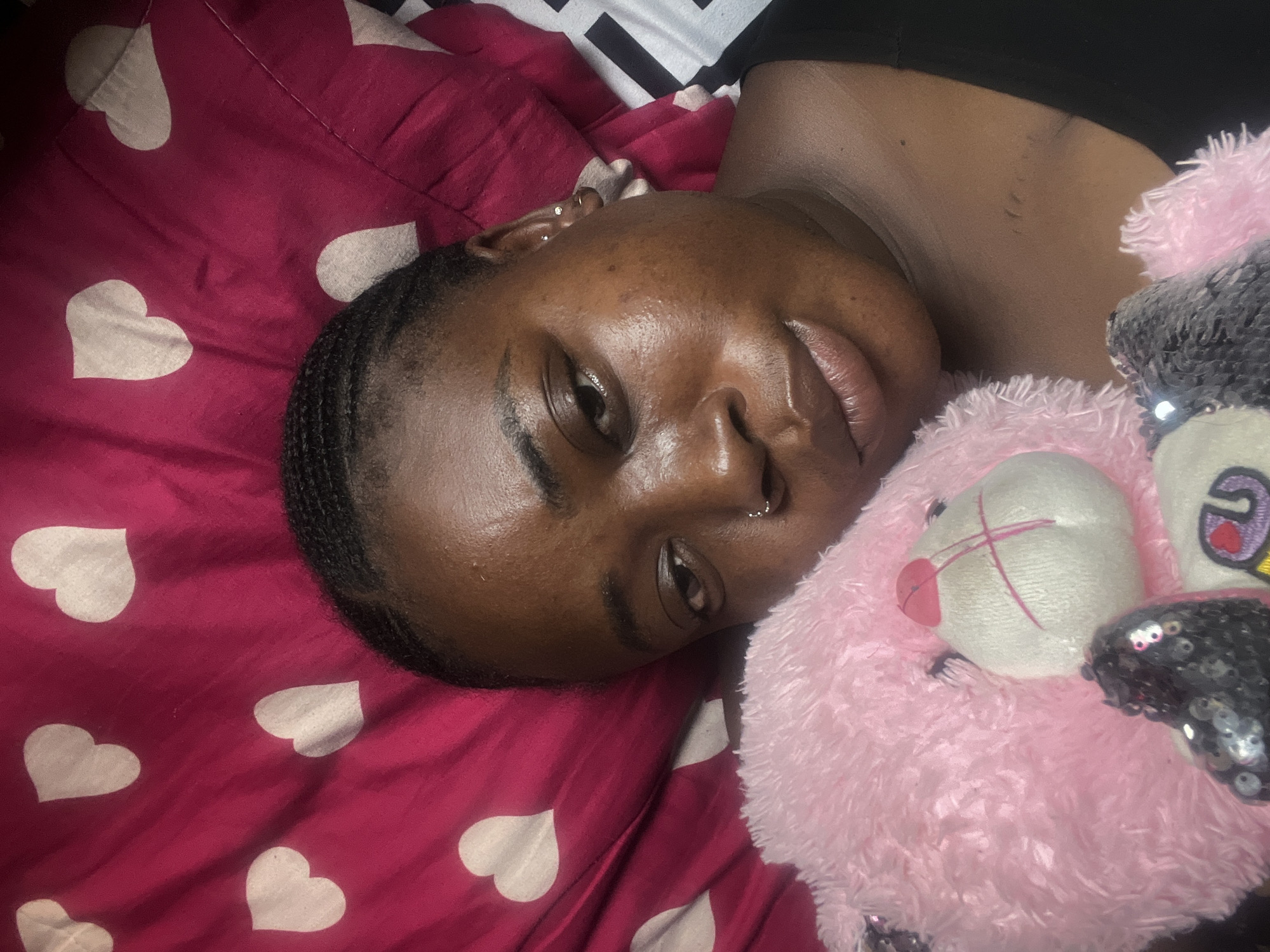 A dark-skinned woman is lying on a red sheet with white hearts and a black and white patterned edge. She has short, dark hair and a nose ring. She is lying next to a pink and white stuffed animal with sequins. The location is not discernible from the image.