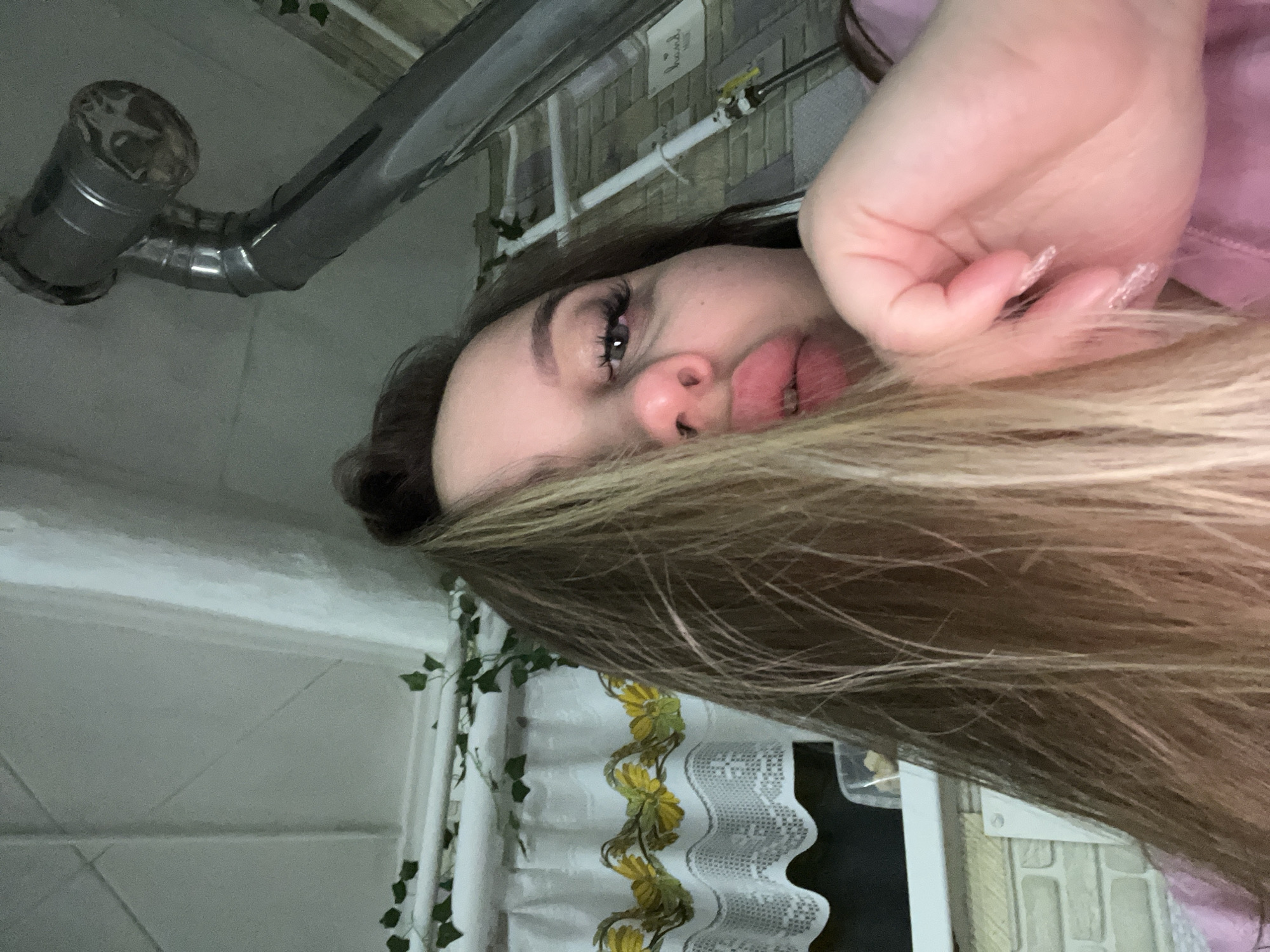 The image shows a young woman in a room with a white-tiled wall and a window covered with a white curtain decorated with yellow flower embroidery and green ivy. She has long brown and blonde hair. She's wearing a pink top. A metal pipe is visible near the ceiling. The location is unknown, but based on the instructions, the city is Port-Katon, Russia.