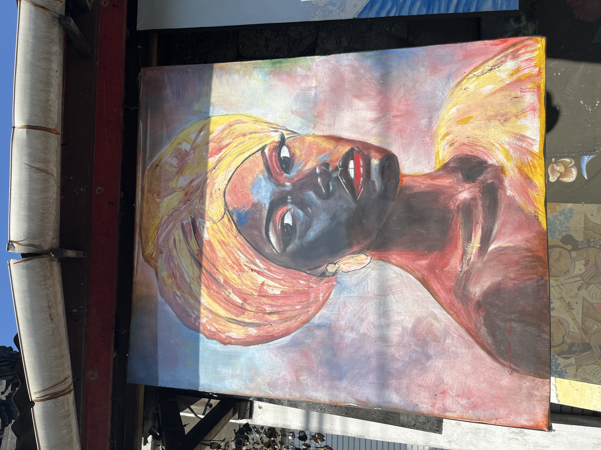 A painting of a woman with dark skin and a yellow head wrap is displayed outdoors. The woman has bright red lips, large eyes with visible eyelashes, and wears a large, golden earring. Her skin is rendered with dark tones, accented by streaks of red and blue. The background of the painting is a soft blend of pink, purple, and blue hues. The painting appears to be leaning against a wall, with other artwork visible below it. In the background, a white gutter and a red beam are visible, suggesting an outdoor setting, possibly a market or art stall, in Sukawati, Indonesia. The sunlight casts shadows on the artwork, indicating it is daytime.