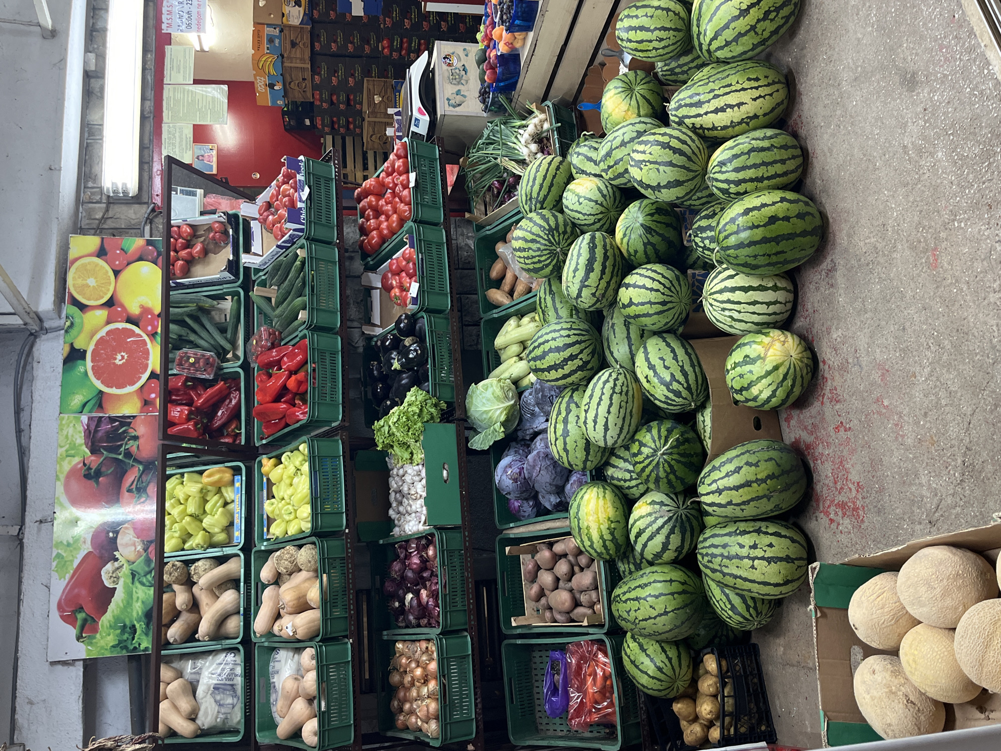 This is a medium shot, eye-level, photograph of a produce stand in Budva, Montenegro. The stand is filled with a variety of fruits and vegetables. In the foreground, there is a large pile of watermelons. Behind the watermelons, there are crates of red cabbage and green cabbage. To the left of the watermelons and cabbages, there are crates of potatoes and carrots. Further back on shelves, there are onions, garlic, lettuce, eggplant, tomatoes, cucumbers, and red peppers. In the background, there is a large poster with images of fruits and vegetables. The floor is concrete. There are no people visible in the photograph.