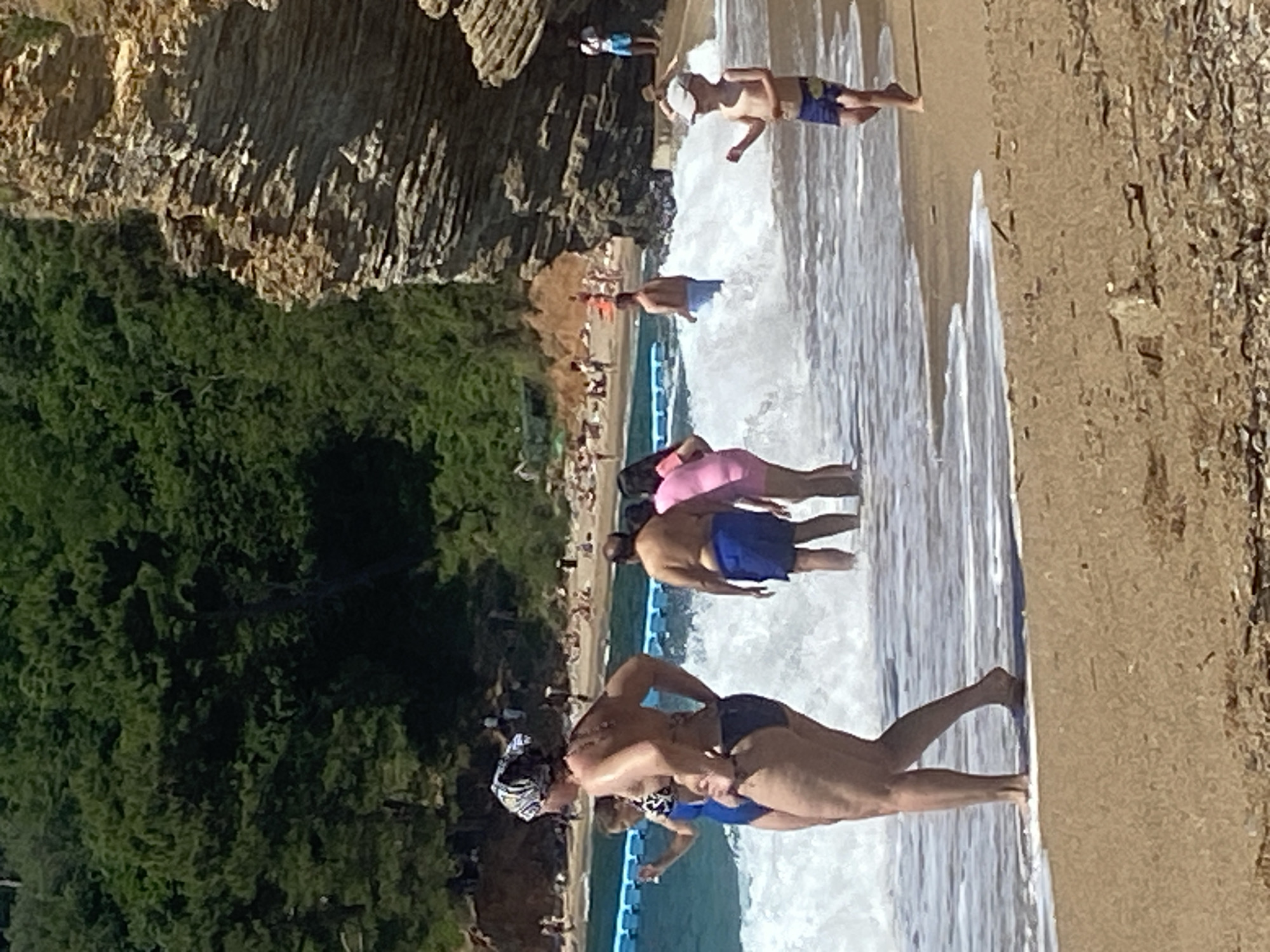 This image depicts a bustling beach scene on a sunny day, consistent with the provided location context of Sveti Stefan, Montenegro. The perspective is from the shallow water, looking towards the sandy shore, rocky cliffs, and verdant vegetation.
In the immediate foreground, several individuals are in the clear, blue-green sea. Two people stand partially submerged; one wears a dark swimsuit and a patterned headscarf, while another is in blue swim trunks. Nearby, a person in a pink swimsuit and blue shorts wades, and a man in blue trunks walks through the gentle white foam of breaking waves.
The midground reveals a sandy beach populated by numerous people. Some individuals are relaxing on towels, while others are strolling along the shore. A prominent blue inflatable barrier or swimming lane extends across the water, separating different areas and containing more bathers.
The background is characterized by a dramatic natural landscape. Steep, rugged rocky cliffs, light in color and partially covered by sparse foliage, rise sharply from the coastline. Below these cliffs, dense green trees and bushes carpet the land, creating a lush contrast with the rock formations. The bright sunlight indicates it is daytime, likely late morning or early afternoon, with clear weather prevailing. The scene conveys a popular coastal destination active with tourists and locals enjoying the sea and sun. No visible text is present in the image.