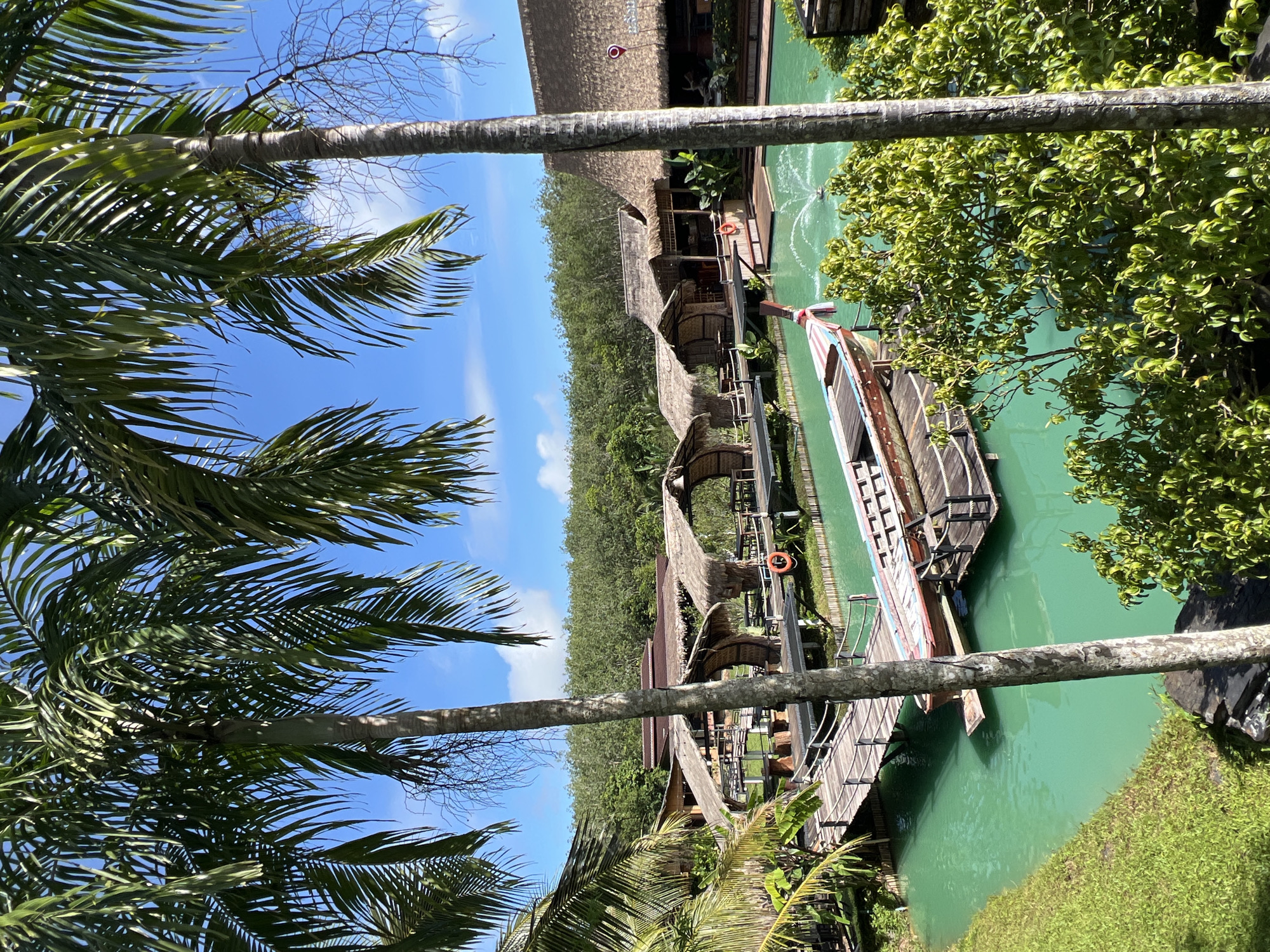 The image depicts a serene scene at a waterfront resort or village, likely in Tambon Pa Kae Bo Hin, Thailand, given the lush tropical vegetation and architecture. The setting is a clear, sunny day, with a bright blue sky and scattered white clouds visible.
The primary subjects are several traditional wooden boats docked alongside a wooden boardwalk and thatched-roof buildings. These buildings appear to be bungalows or shops, constructed with natural materials, suggesting a rustic and eco-friendly aesthetic. The water is a vibrant, emerald green, hinting at its clarity or the presence of algae, and a fountain is visible in the distance, adding to the tranquil atmosphere.
Palm fronds frame the top and left sides of the image, emphasizing the tropical location. The boardwalk is made of weathered wood and leads to the various structures. Life preservers are visible on the boardwalk and near the water, indicating safety precautions.
While no people are clearly visible in the image, the presence of the boats, boardwalk, and buildings implies human activity and habitation. The overall mood is peaceful and relaxing, typical of a tropical getaway. The time of day appears to be midday, judging by the bright sunlight and the shadows cast. There is no visible text in the image.
