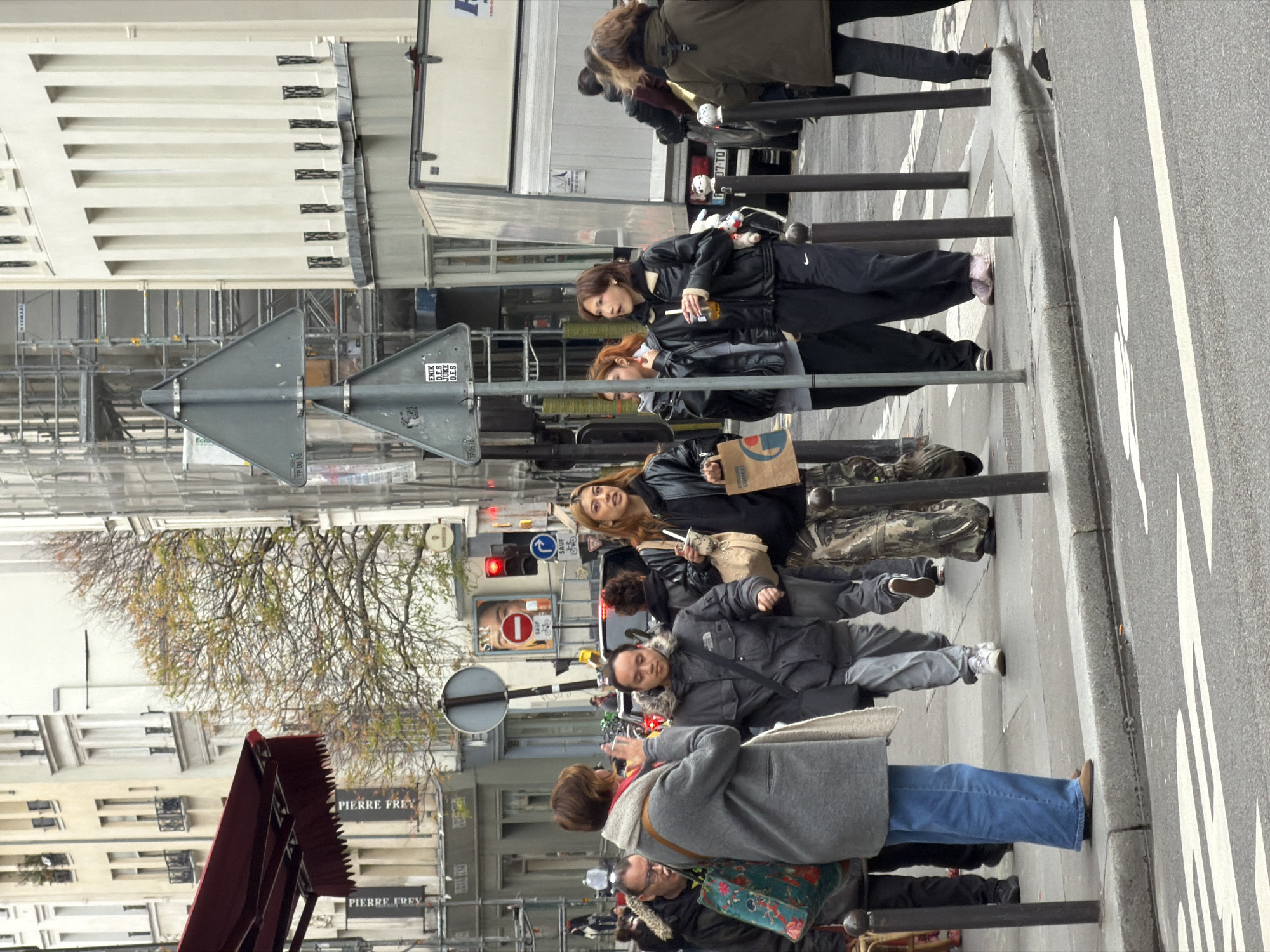 The image captures a street scene in Paris, France, on an overcast day. Pedestrians are visible on a grey sidewalk, with a paved road and white crosswalk markings in the foreground.
In the immediate left foreground, a woman with short brown hair, wearing a grey coat, light-colored scarf, and blue jeans, stands with her back mostly to the viewer, holding a silver smartphone up as if taking a photograph. To her left, partially obscured, a man with glasses in a dark jacket carries a teal-colored tote bag with a floral pattern.
In the middle ground, moving from left to right along the sidewalk, several individuals are present. A man in a dark grey, fur-lined hooded jacket with a black messenger bag walks with a determined expression. To his right, a woman with long light brown hair, dressed in a black leather jacket and camouflage-patterned pants, holds a light-colored handbag and a brown paper shopping bag, looking towards her right. Next to her, a woman with short brown hair, wearing a black leather jacket and black track pants with a white Nike logo, carries a clear plastic cup with a straw, also looking to her right. Behind her, another woman in a black leather jacket is partially visible. Further right, a person in an olive green hooded jacket has their back to the viewer.
Street furniture includes multiple dark grey bollards lining the sidewalk's edge. A metal pole supports two triangular grey warning signs, the lower one featuring an "ENIK O.E.S." sticker. In the background, a red traffic light is active, alongside a circular red 'no entry' sign with a bicycle symbol, and a blue circular sign indicating a mandatory right turn 'SAUF VÉLO' (except bicycles).
The urban environment features multi-story Parisian buildings. On the left, a dark red awning extends over a shop entrance, with "PIERRE FREY" visible on signs below. A building on the right exhibits a uniform facade with vertical decorative elements and small windows with bars. Scaffolding is visible on a building further in the background. Bare-branched trees with sparse yellow-green foliage are present along the street. A large white truck is parked partially visible on the right side of the road.