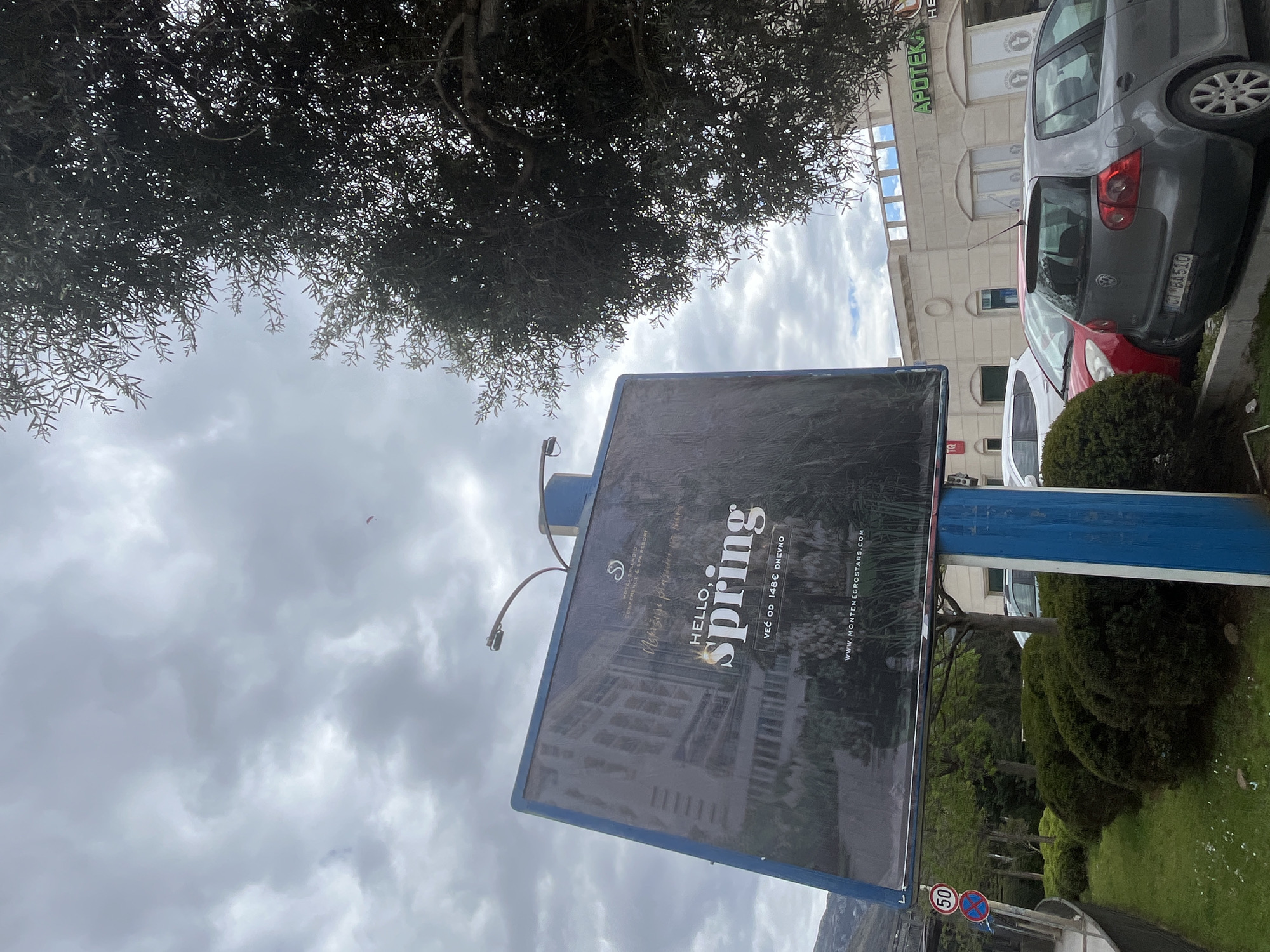 The image shows a billboard advertising "HELLO, spring" for Hotel Splendor Conference & Spa Resort, located in Boreti, Montenegro, under a cloudy sky partially obscured by a tree. The billboard is positioned on a blue support structure. To the right of the billboard are parked cars, a building with "APOTEKA" visible, and manicured greenery. A "50" speed limit sign is also visible in the lower-left. The license plate on the grey car is CBA510.