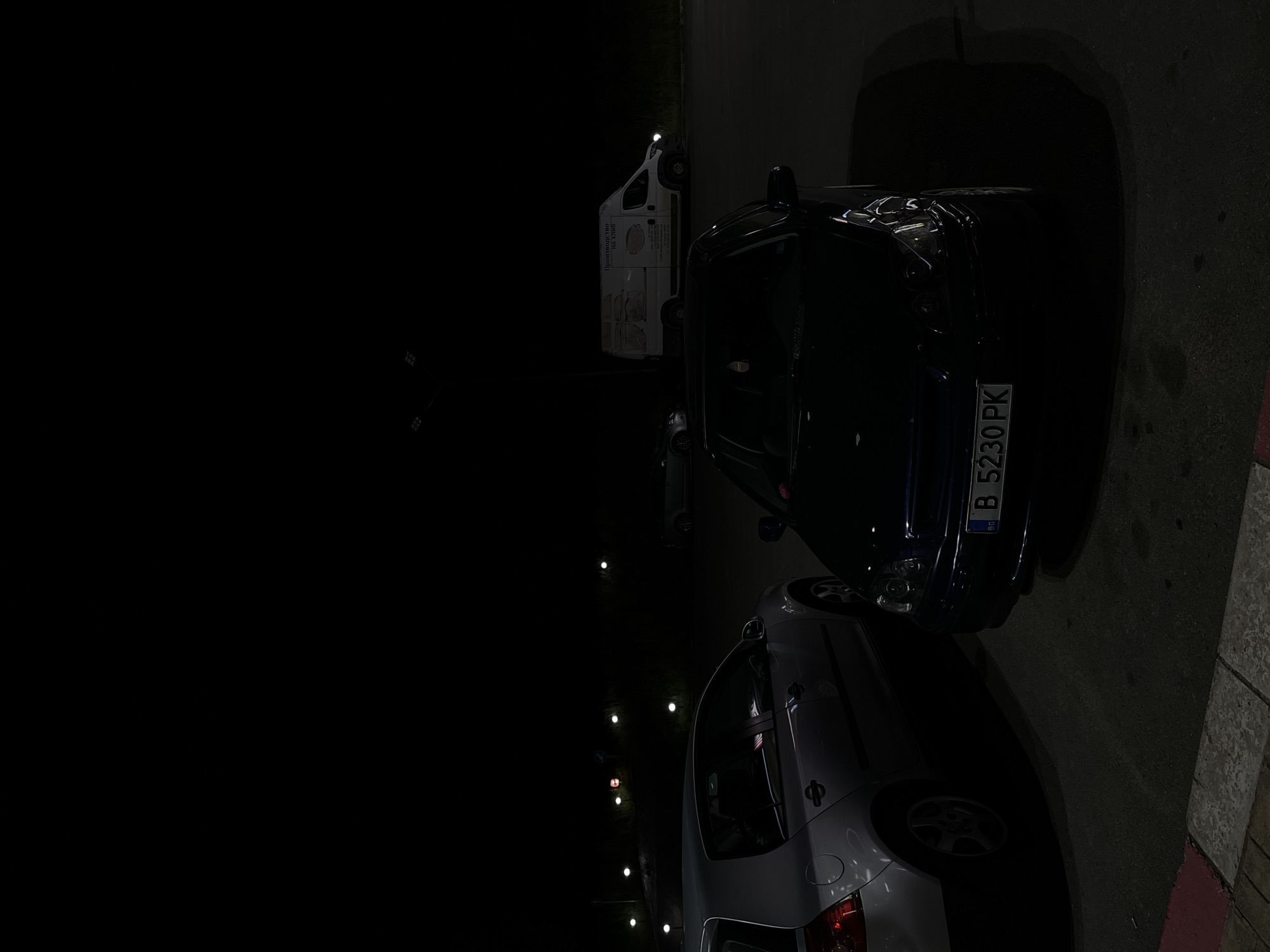 The image depicts a dimly lit street at night, possibly in or near Yabalchevo, Bulgaria. Two cars are parked on the right side of the frame. The closer car is light-colored. Behind it is a darker-colored car with a license plate visible, "B 5230 PK," suggesting the car is registered in Bulgaria. A white van with some writing on its side is parked further in the background. The street is dark, with only faint street lights visible. The environment appears urban, and the scene is stationary.