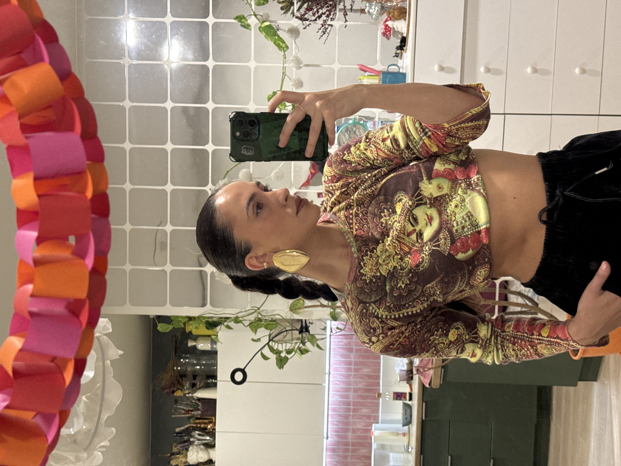 A woman is taking a selfie in front of a mirror decorated with a colorful paper chain. She is wearing a patterned crop top, black pants, and large gold earrings. She holds a smartphone in her left hand. The background includes a kitchen with white cabinets, a tiled wall, and various plants and decorations on the shelves. The location is possibly a domestic interior, but there is no explicit information to confirm this or its location within Krasnoyarsk, Russia.