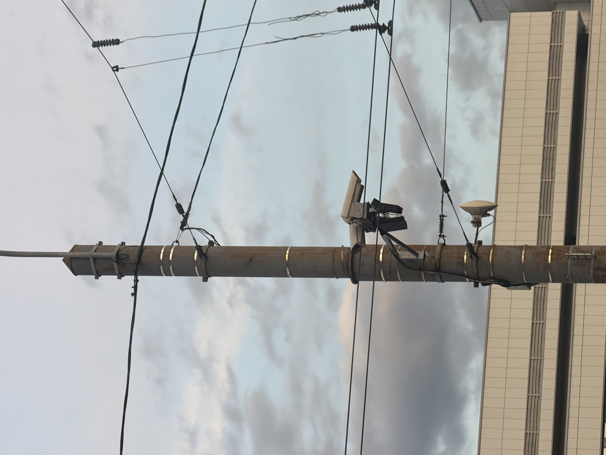 A metallic utility pole stands vertically in the center of the frame, extending from the bottom edge towards the top. It is adorned with several horizontal bands and metallic attachments. Attached to the pole at different heights are various electrical wires and two security cameras. One camera, white and boxy, is mounted on a bracket and faces left, with another partially visible camera directly below it. Lower down the pole, a dish-shaped antenna is also mounted. Multiple power lines crisscross the sky behind the pole. In the background, a light-colored building with horizontal paneling and horizontal vents is visible. The sky is cloudy with patches of blue.