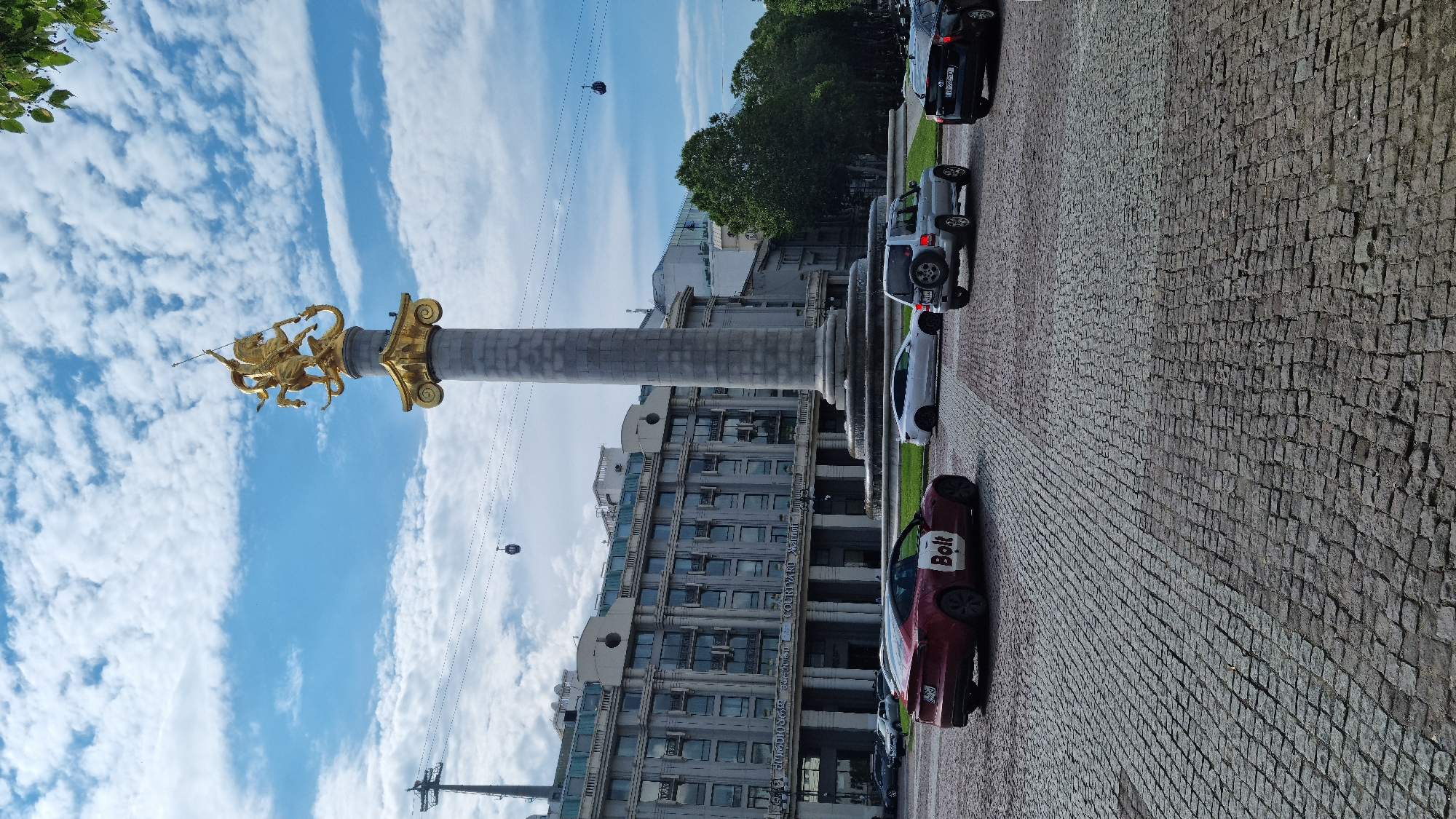 The image captures a street scene in Tbilisi, Georgia, with a prominent monument in the background and vehicles in the foreground. The monument, topped with a golden sculpture of a horseman, stands tall on a stone column, casting a shadow on the paved street. The building behind the monument appears to be a hotel, with multiple stories and arched windows. A red car with "Bolt" branding is parked on the right side of the image, alongside a silver SUV and a black car. The street is paved with cobblestones, and the sky is filled with white clouds. Cable cars can be seen suspended in the air, indicating a popular tourist area.