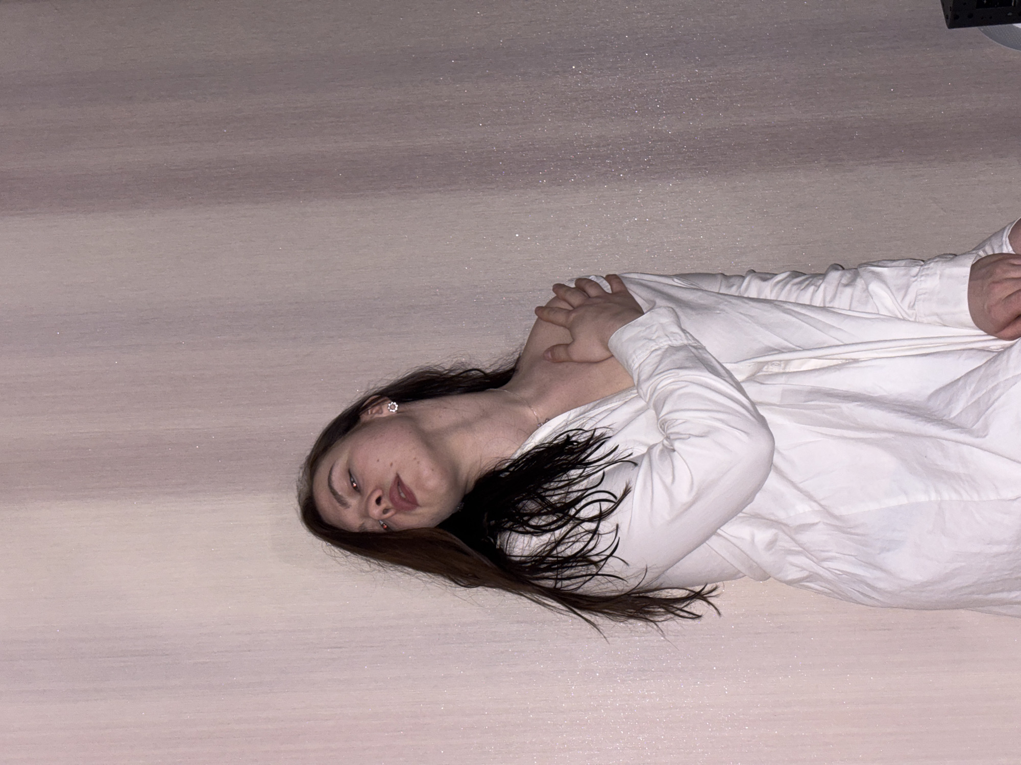 The image displays a young woman lying down on a light-colored, possibly white or pale gray, textured surface that exhibits a subtle glitter or sparkle. Her body is oriented horizontally across the frame, with her head facing the left. She possesses long, dark brown hair that cascades around her shoulders and back. Her complexion is fair, and faint freckles are visible across her nose and cheeks.
She is dressed in a white or light-toned long-sleeved button-up shirt or blouse. Her arms are crossed over her midsection, suggesting a relaxed or introspective pose. The woman is gazing directly at the viewer with a neutral, possibly pensive, expression. A small, ornate earring, resembling a silver flower, is visible in her left ear. A reddish reflection is present in her eyes, likely a result of the camera flash or artificial lighting conditions. The background is simple and uniform, composed entirely of the textured surface she rests upon. An indistinct dark object is partially visible in the top right corner, which could be part of a fixture or furniture. The setting is indoors, and the image was captured in Perm, Russia. There is no visible text in the image.