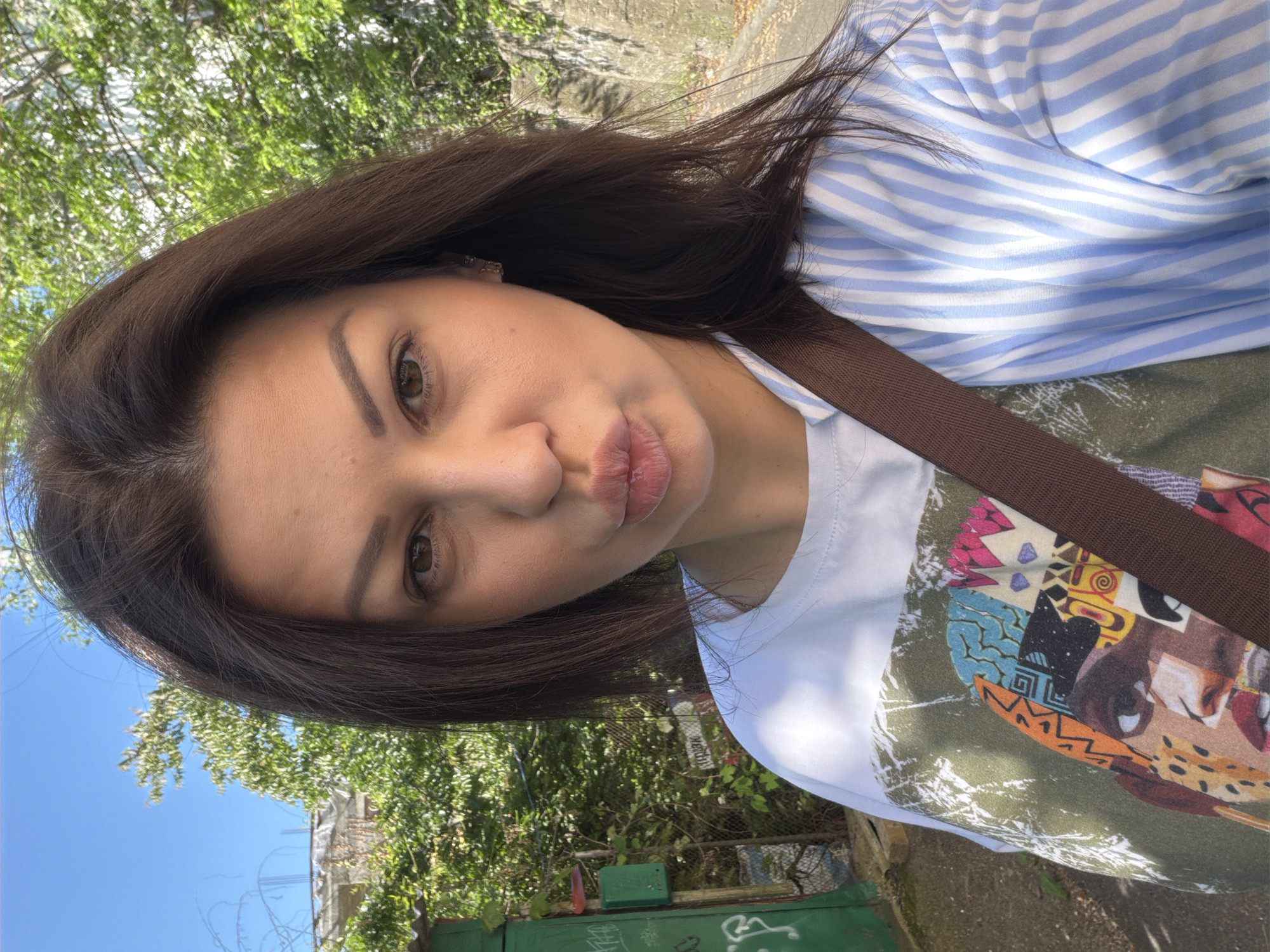 An outdoor selfie captures a woman on a sunny day in Sochi, Russia. The woman, with dark brown hair and brown eyes, faces the camera with pursed lips. She is wearing a white t-shirt with a vibrant, multi-colored graphic print of a stylized face, partially covered by a light blue and white striped shirt. A brown strap, likely from a bag, extends diagonally across her chest. A light-colored earring is visible in her right ear. The background to the left features a green metal fence, a green utility box with graffiti, and dense green foliage, under a clear blue sky. Partially visible structures or distant buildings are present above the fence line. To the right, a path is bordered by a concrete or stone wall covered with sparse vegetation, behind which additional green trees and bushes are visible.