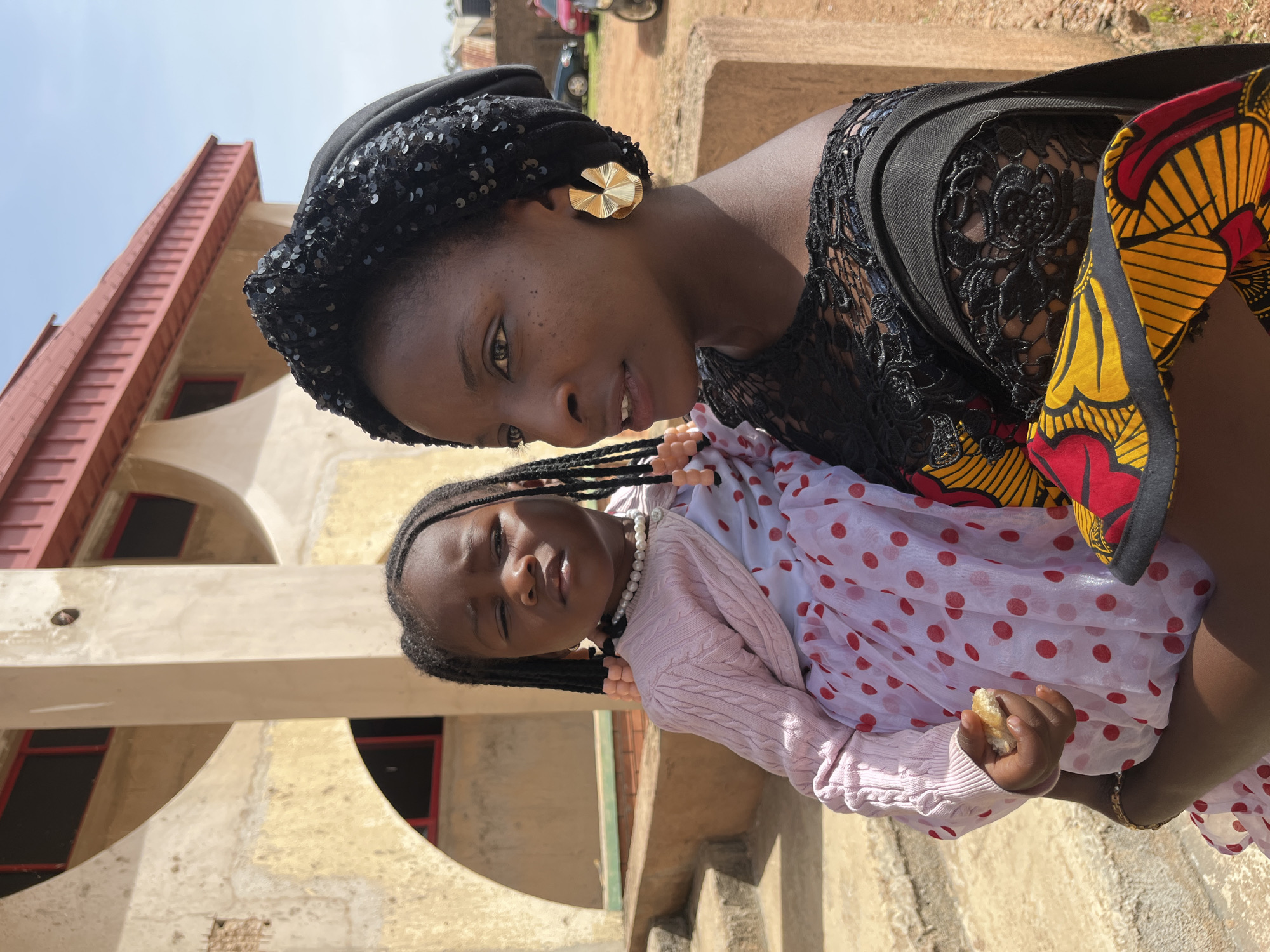 In the city of Bukuru, Nigeria, a young woman wearing a black sequined headscarf and an elaborate black lace top holds a small child. The child, with dark braided hair adorned with pink beads, wears a pink knit sweater over a white dress patterned with red polka dots. The child's right hand holds a piece of bread. The woman is wearing large, gold, fan-shaped earrings. Behind them, a building with arched windows and a red roof is visible. The scene is outdoors, with a clear blue sky overhead. The ground appears to be paved with concrete or stone, and there are visible steps leading up to the building. In the background, other buildings and vehicles are faintly visible.